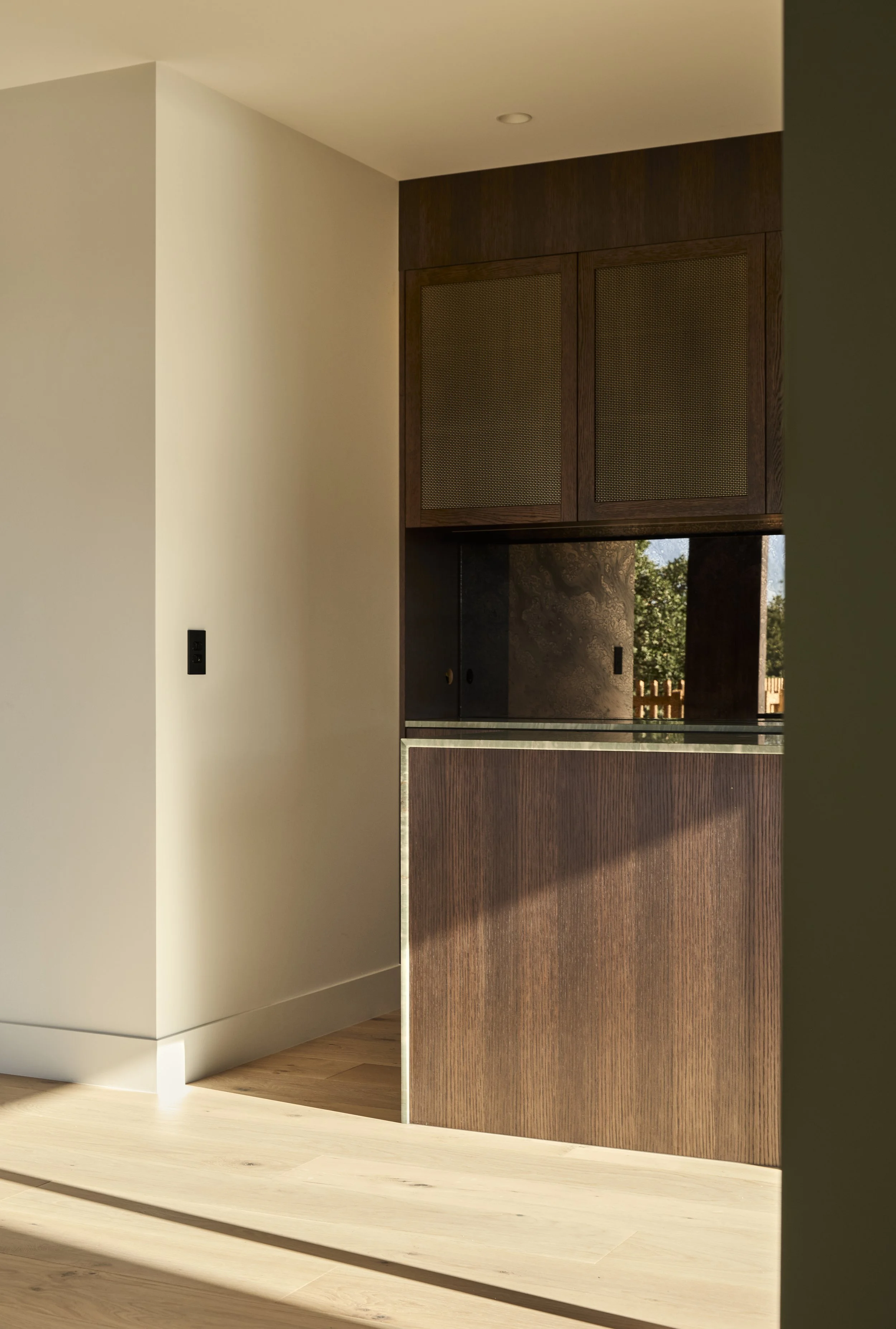 Interior view of a modern room showing a white wall and a partial view of a wooden cabinet with glass window, sunlight on light-colored wooden flooring.