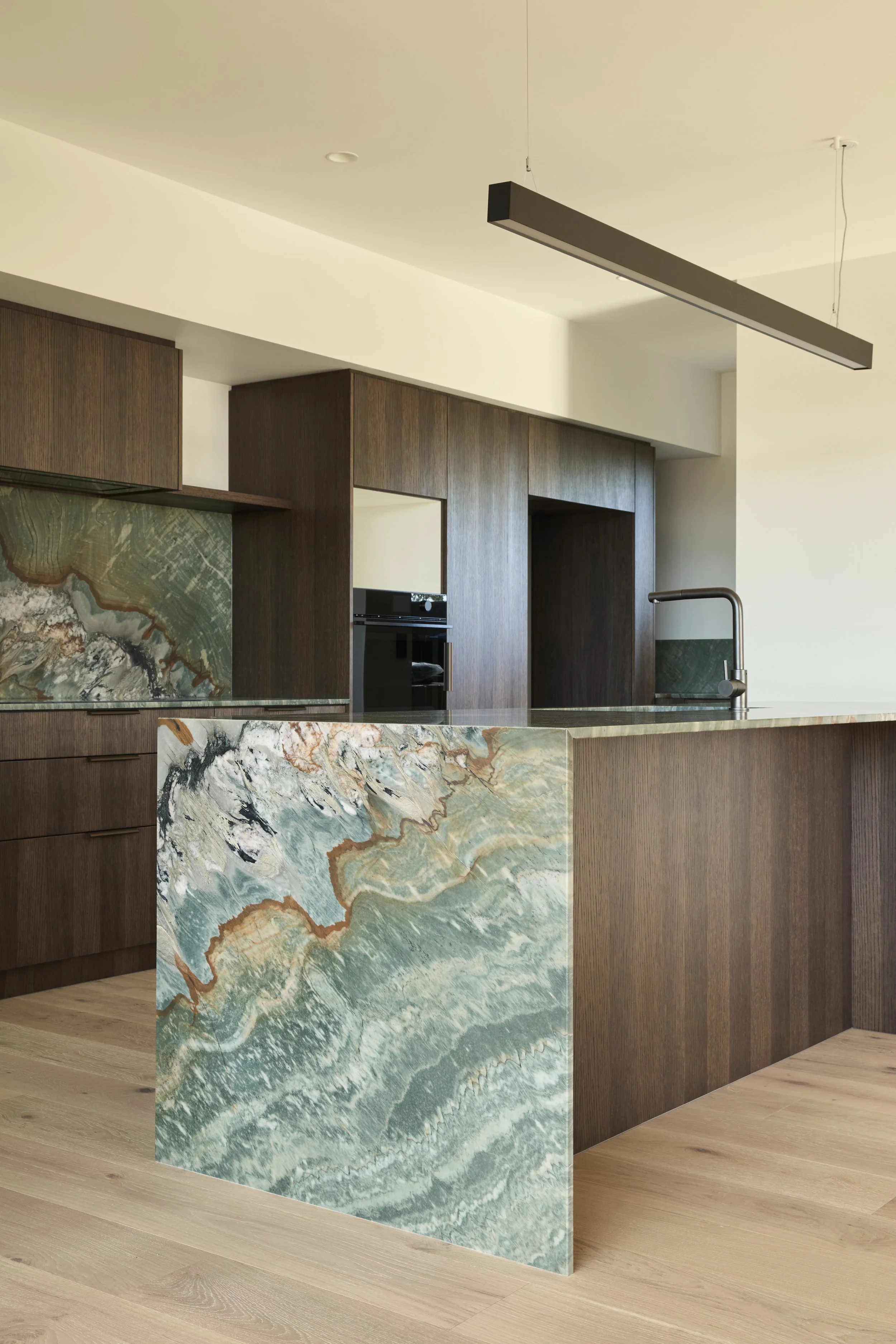 Modern kitchen with dark wood cabinets, a marble island with green, brown, and white veining, and a sleek black faucet. Minimalist design with natural light.