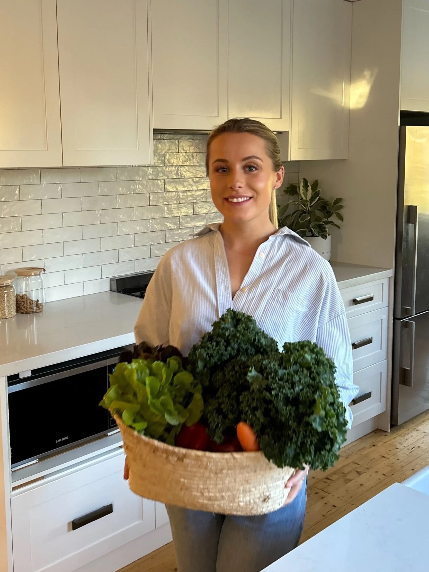 A woman standing in a kitchen holding a basket of fresh vegetables, including kale, leafy greens, and carrots, smiling at the camera.