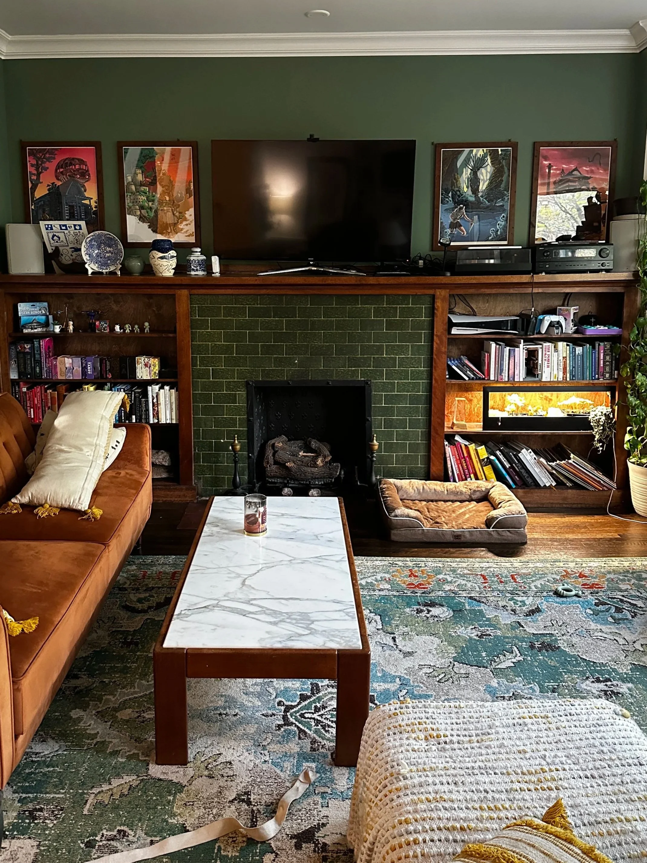 Living room with a green wall, a black TV, framed artwork, a fireplace with a green brick surround, a wooden bookshelf filled with books and decorative items, a sofa with a pillow, a coffee table with a candle, a dog bed, and a patterned area rug.