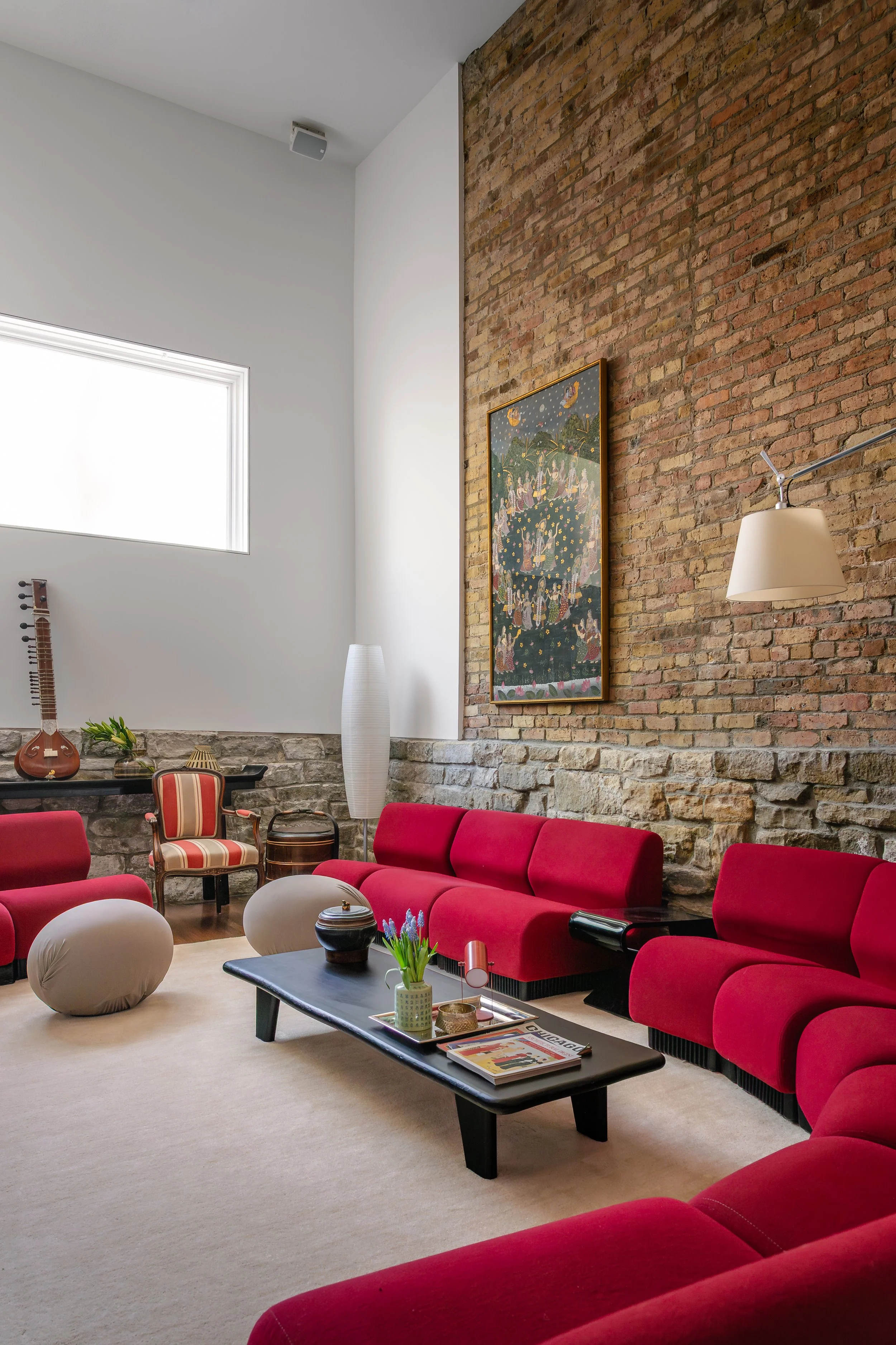 Living room with red sofas, a black coffee table, modern floor lamp, a framed Asian-inspired artwork on an exposed brick wall, decorative vases, and a stone fireplace with musical instruments and plants near a large window.