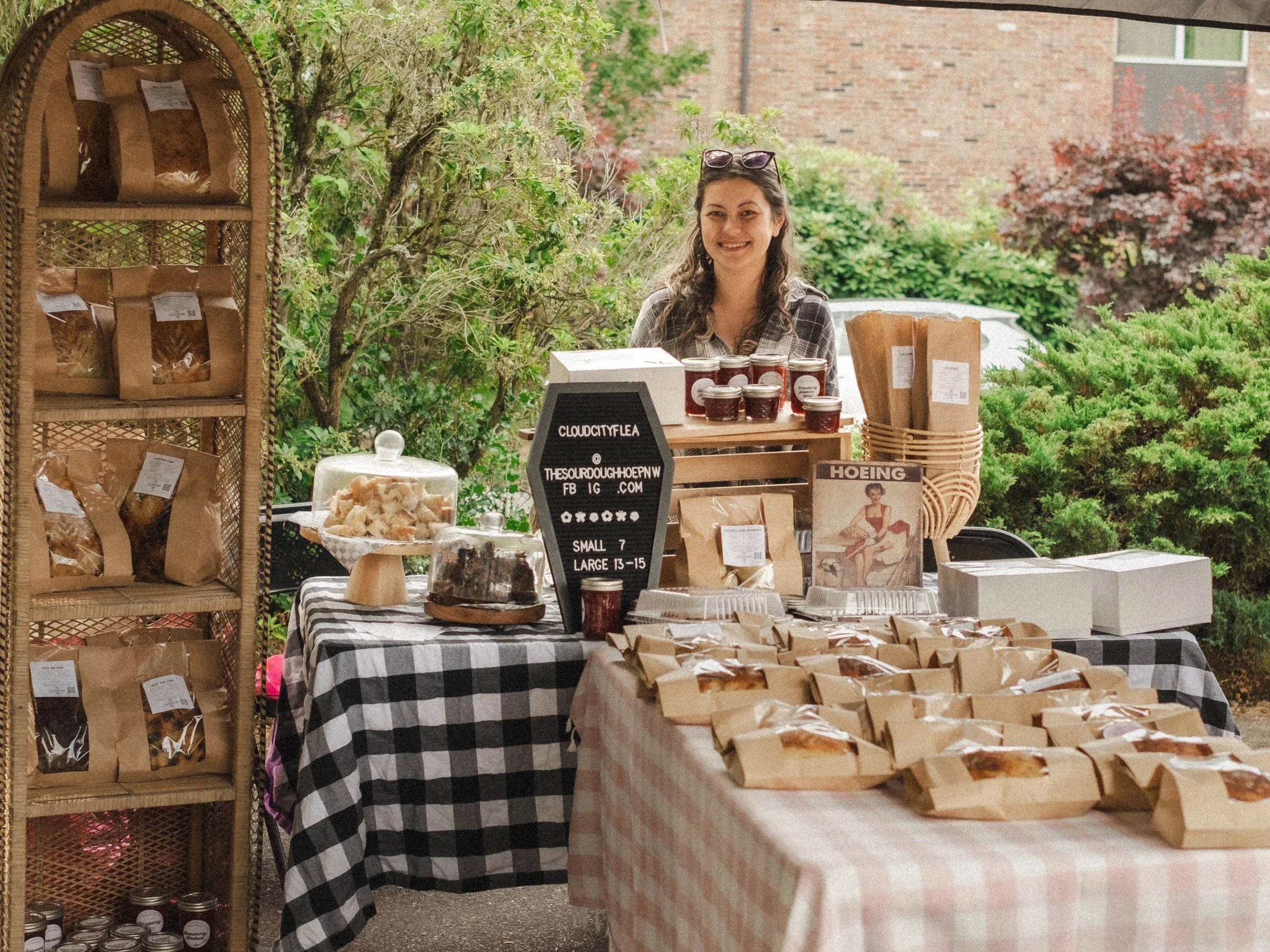 A woman with sunglasses on her head standing behind a table at an outdoor market stall, with jars of jam, baked goods, and packaged goods displayed on tables and shelves, with greenery and trees in the background.