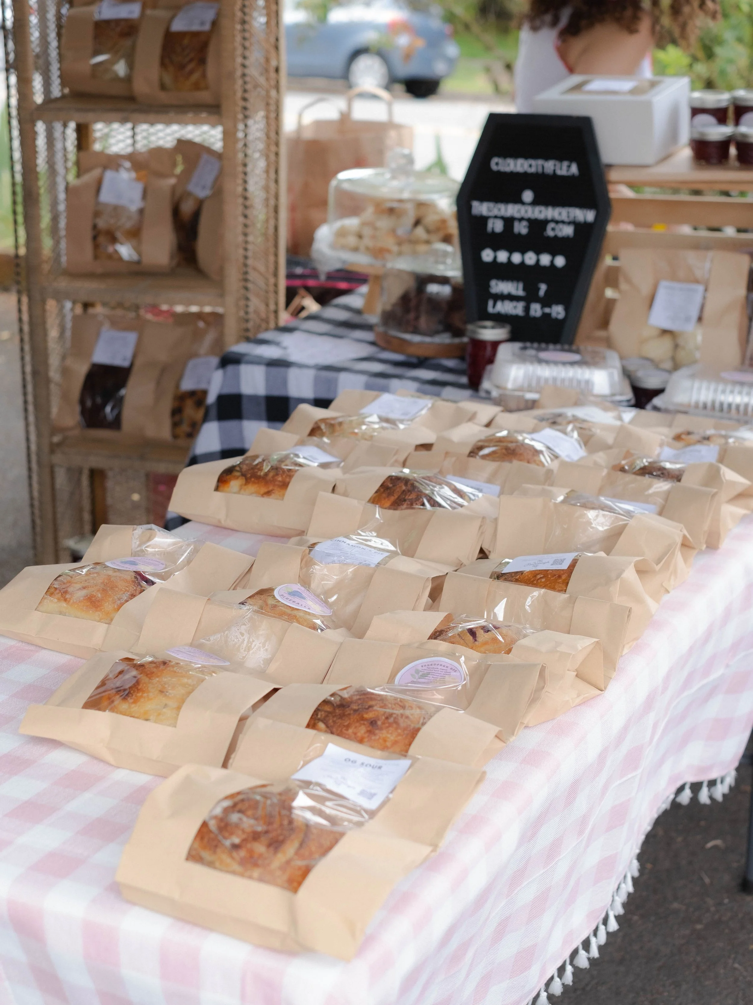A table covered with a pink and white checkered tablecloth displays individually wrapped baked goods at an outdoor market. Behind the table, a chalkboard sign provides product details. Shelves on the side hold more baked items in paper bags.