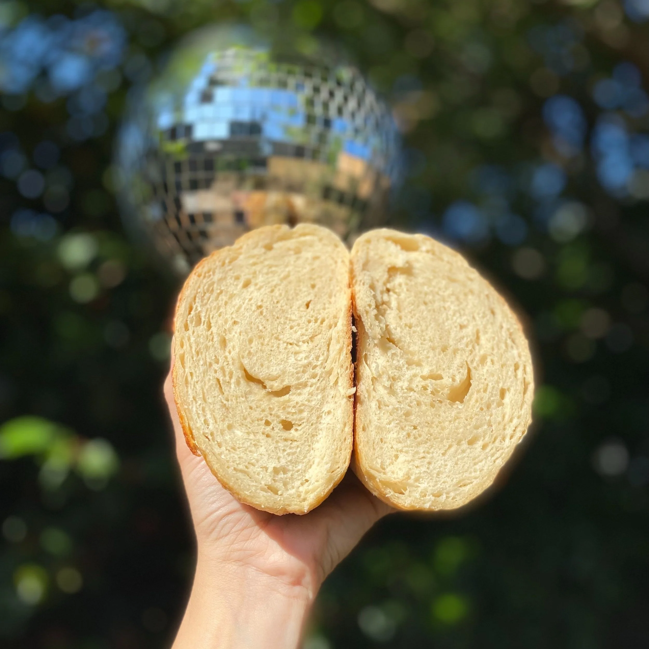 A hand holding a halved beige bread roll with a disco ball in the background outdoors.