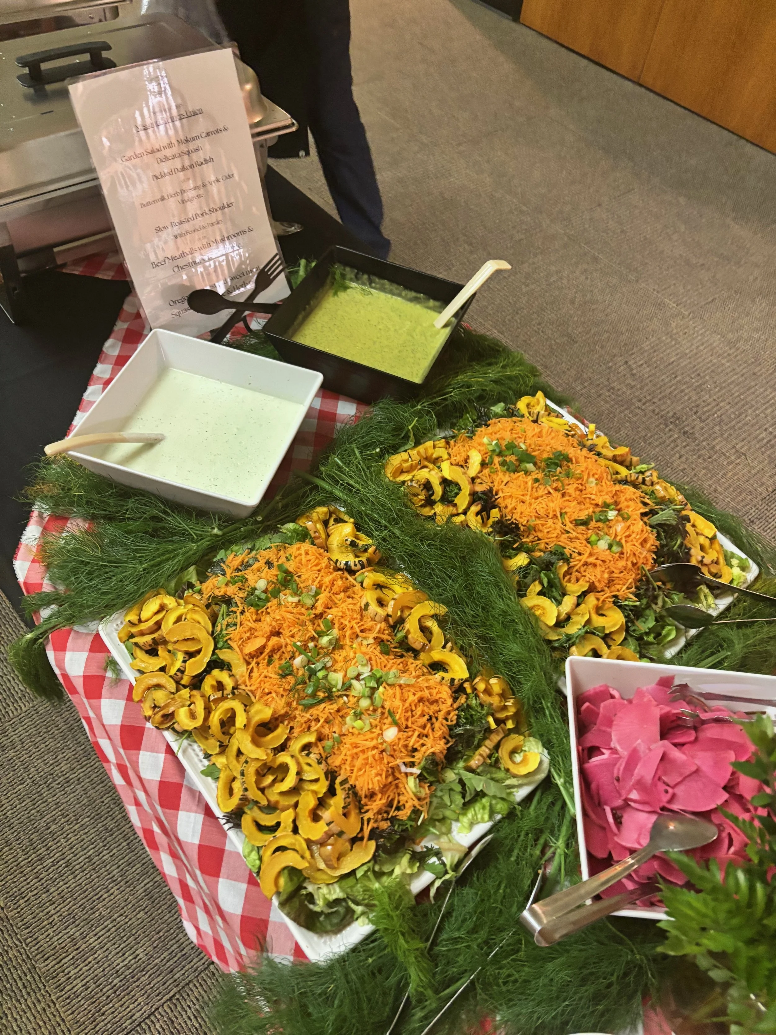 Salad bar with yellow squash, shredded carrots, leafy greens, pink pickled vegetables, and dressings in bowls, on a table with a red checkered tablecloth.