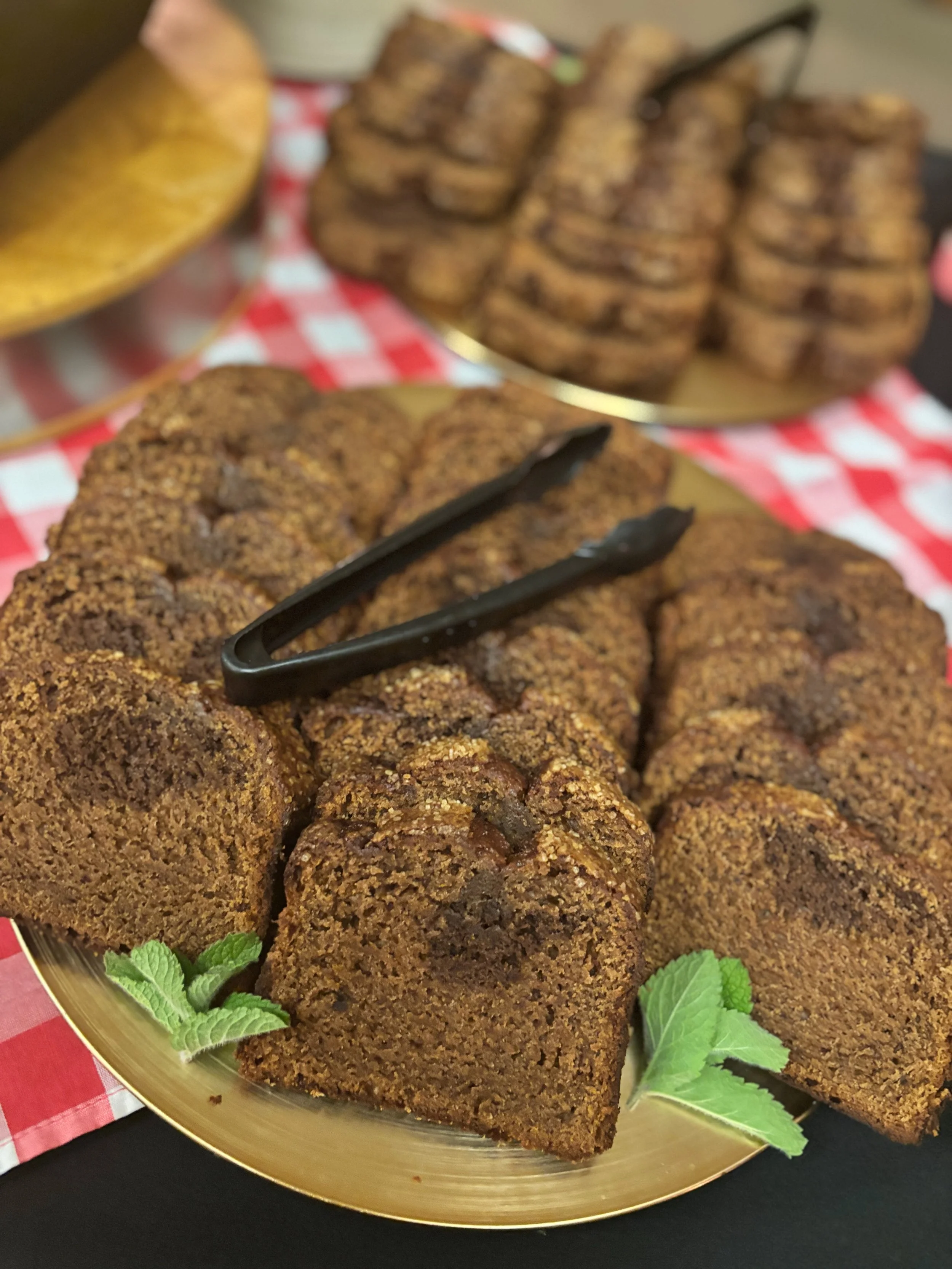 Plate of sliced banana bread with a black tongs on top, garnished with mint leaves, with more slices of banana bread in the background on a checkered red and white tablecloth.