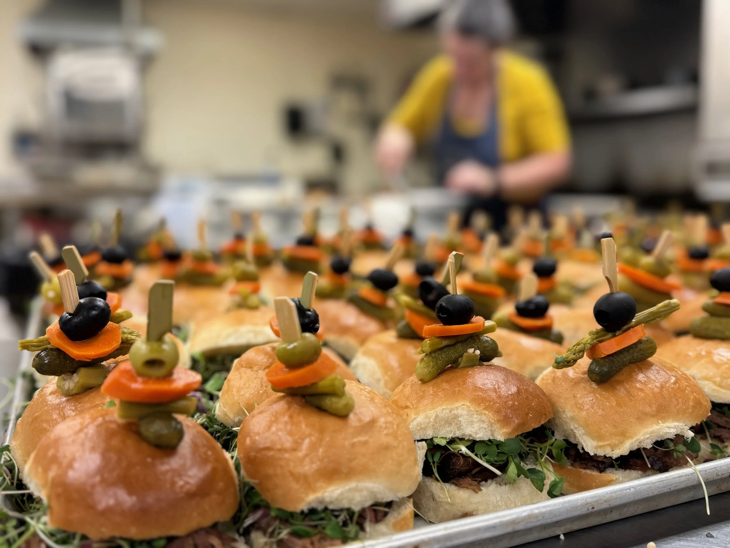Tray of gourmet sliders topped with mixed vegetables and black olives, in a professional kitchen with a blurred chef in the background.