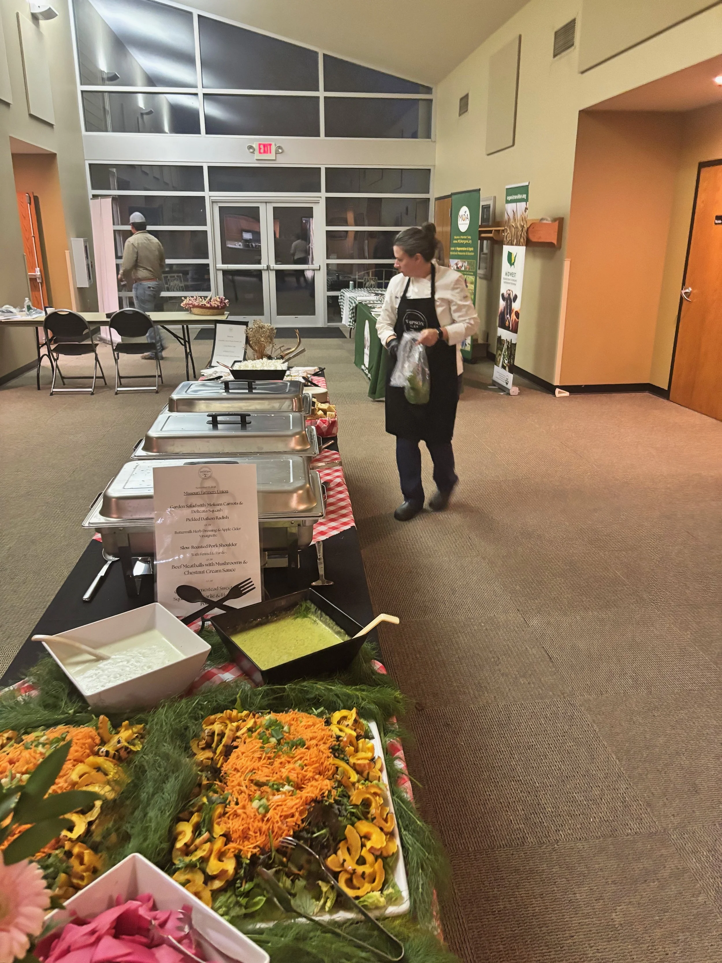 A woman selecting food from a buffet table at an indoor event space with salad, soup, and other dishes.