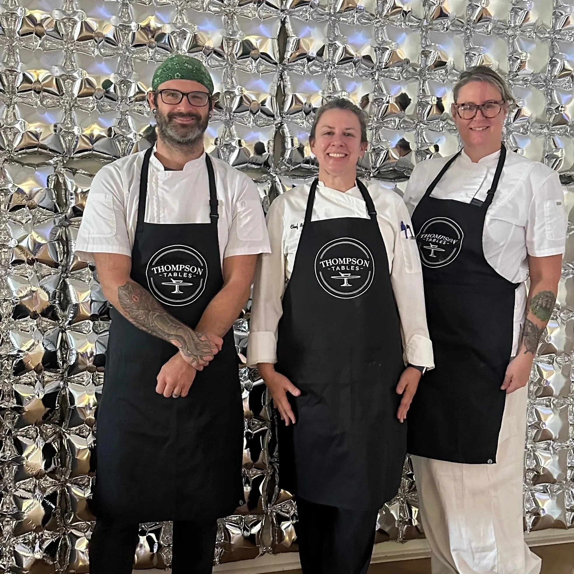 Three chefs standing side by side wearing black aprons with the logo "Thompson Tables" in front of a metallic, reflective backdrop.