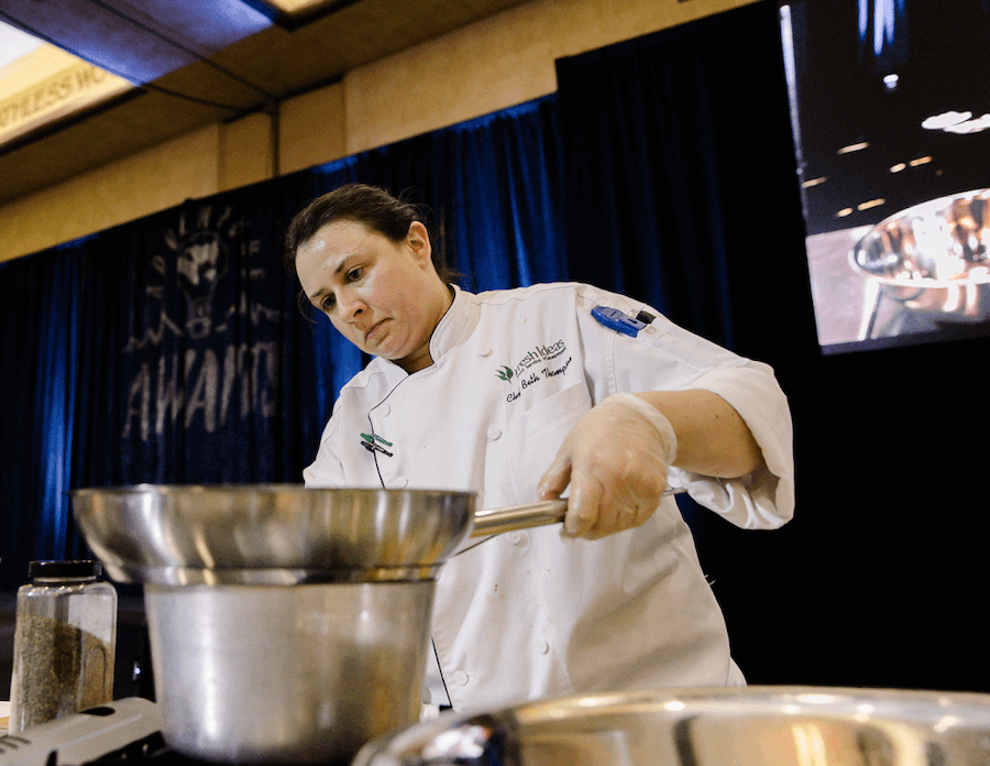 A female chef wearing a white chef's coat and gloves is cooking with a frying pan on a stove during a demonstration or cooking event.
