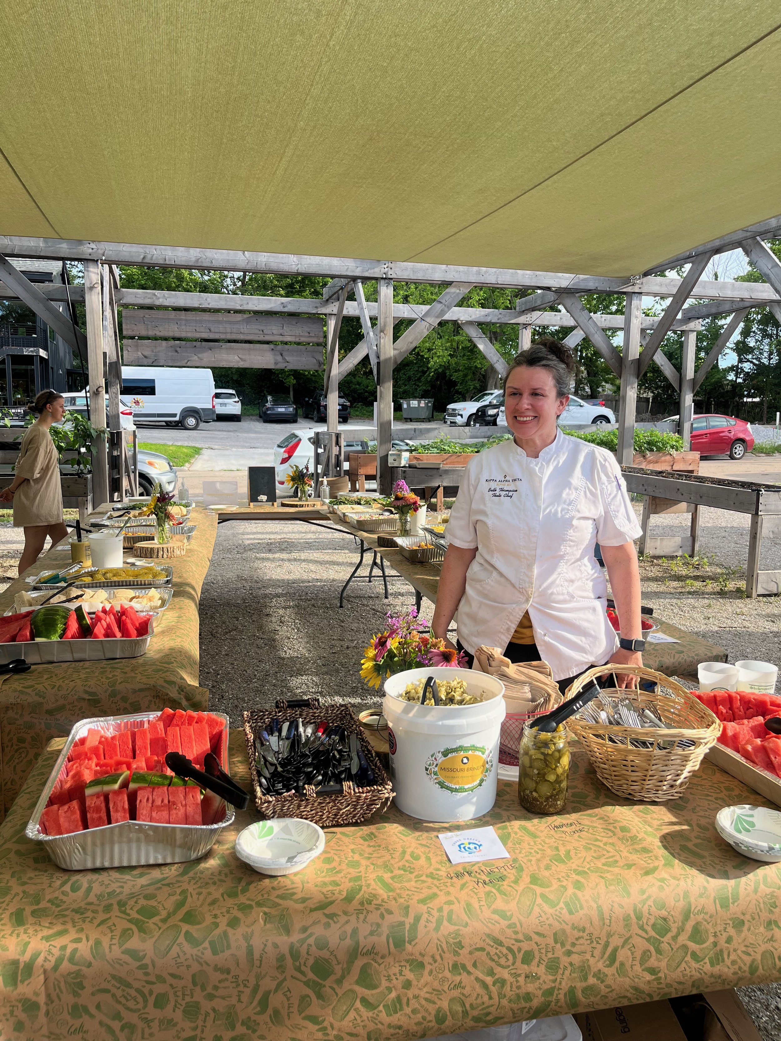 A smiling woman in a chef's coat standing behind a table with watermelon slices and other food items under an outdoor canopy.