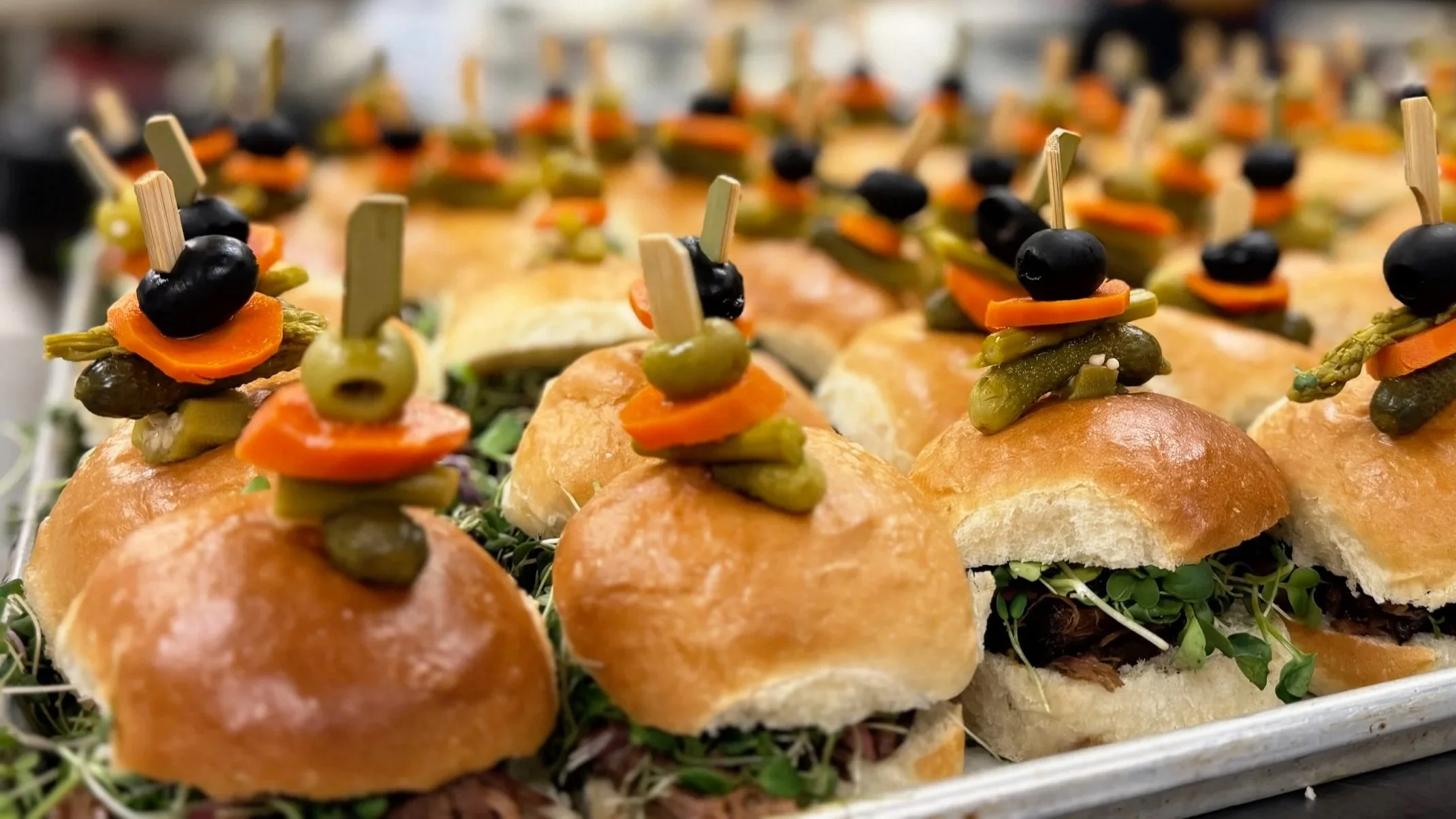 Mini sandwiches with various toppings, topped with toothpicks holding black and green olives, placed on a tray with greens.