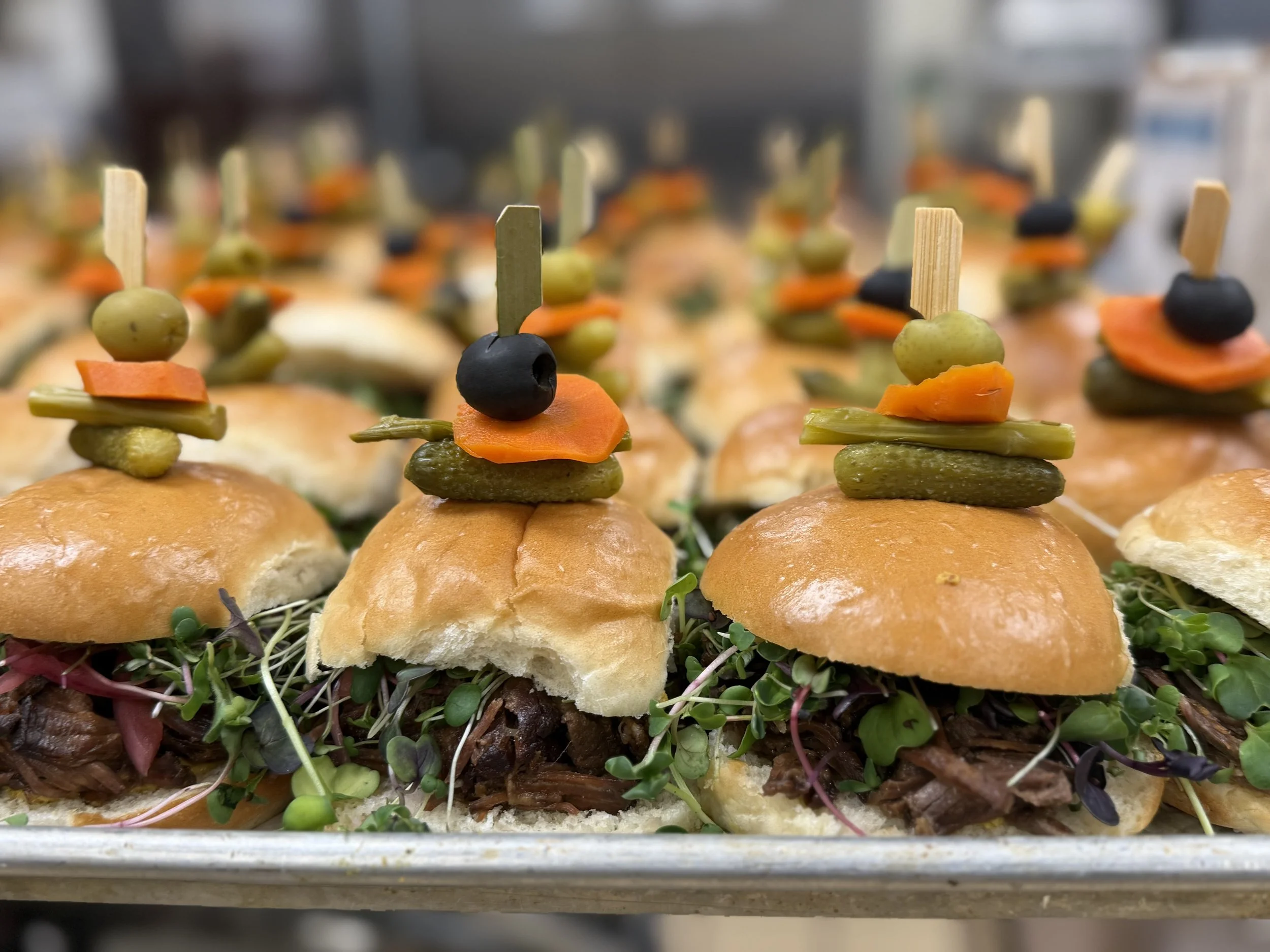Mini sandwiches with shredded beef, microgreens, and small buns topped with skewers of green olives, carrots, green beans, and black olives.