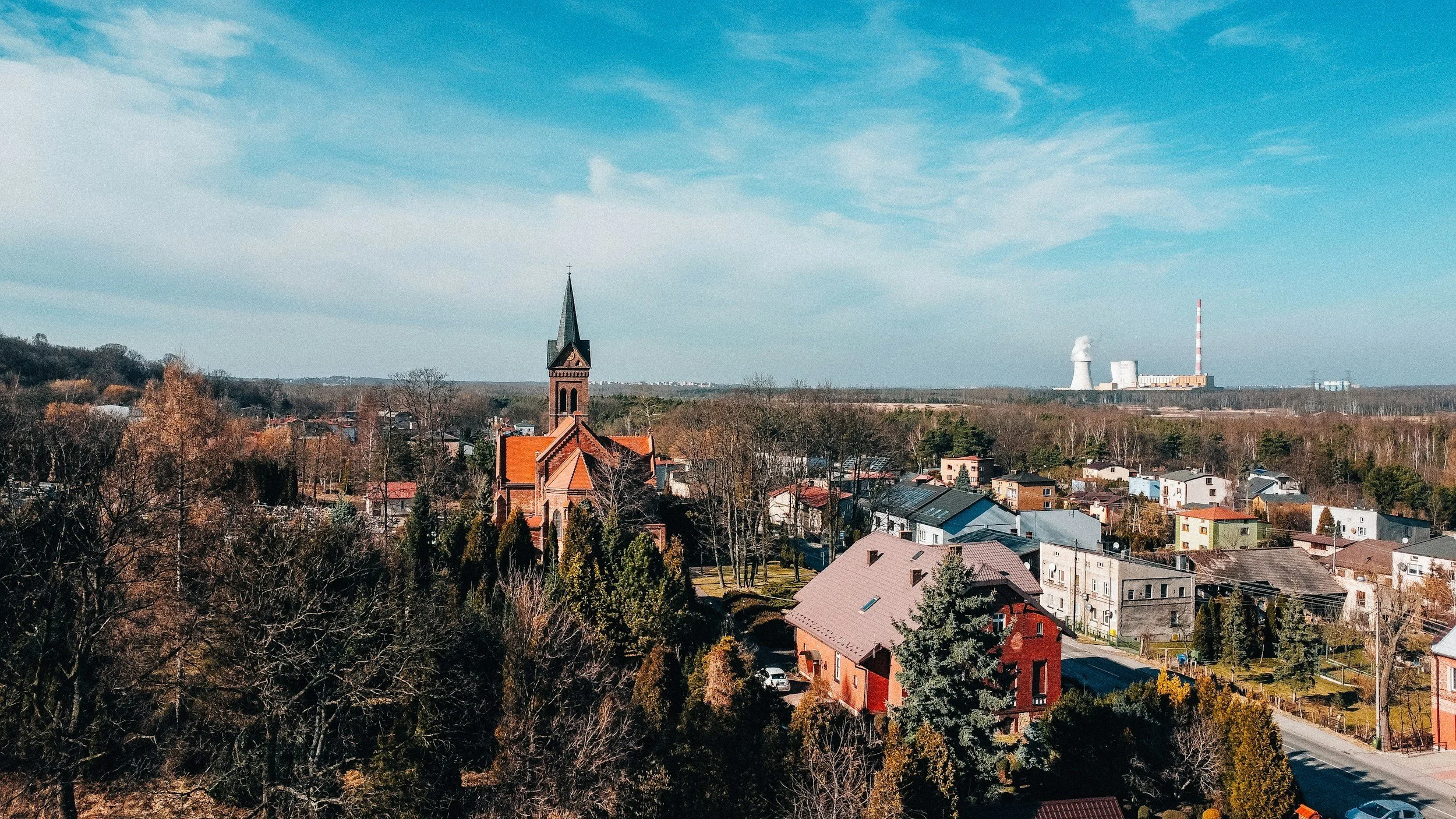 A small town with a church in the foreground, surrounded by trees and residential houses, with an industrial plant in the background under a blue sky.