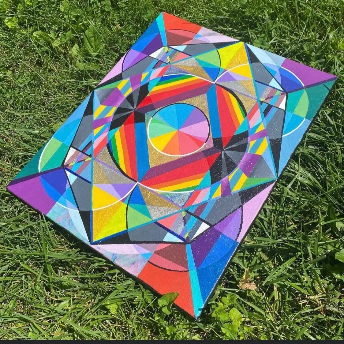 A colorful geometric painting with circles, triangles, and sharp lines on a canvas placed on grass outdoors.