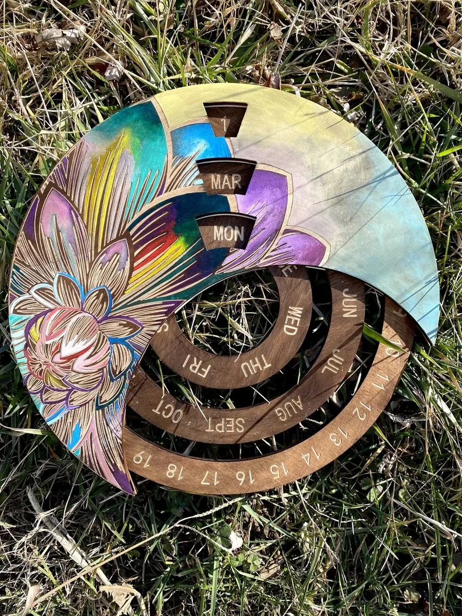 Colorful circular calendar with rings displaying months and days, featuring decorative floral and abstract painted design, placed on grass.