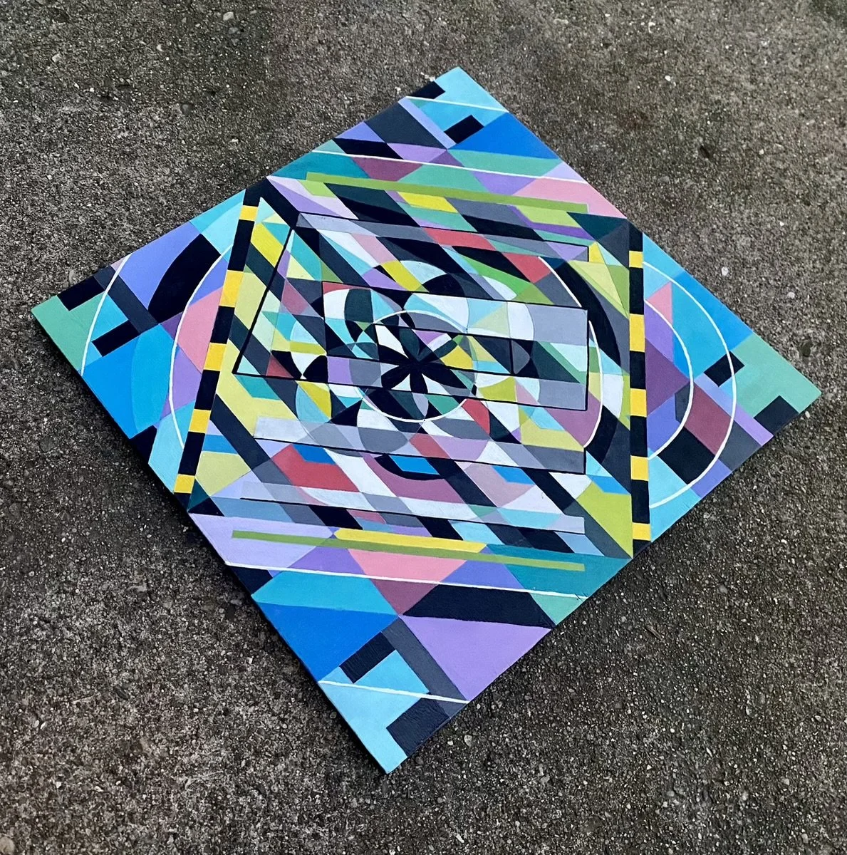 Colorful geometric abstract painting on a canvas placed on a rough asphalt surface.