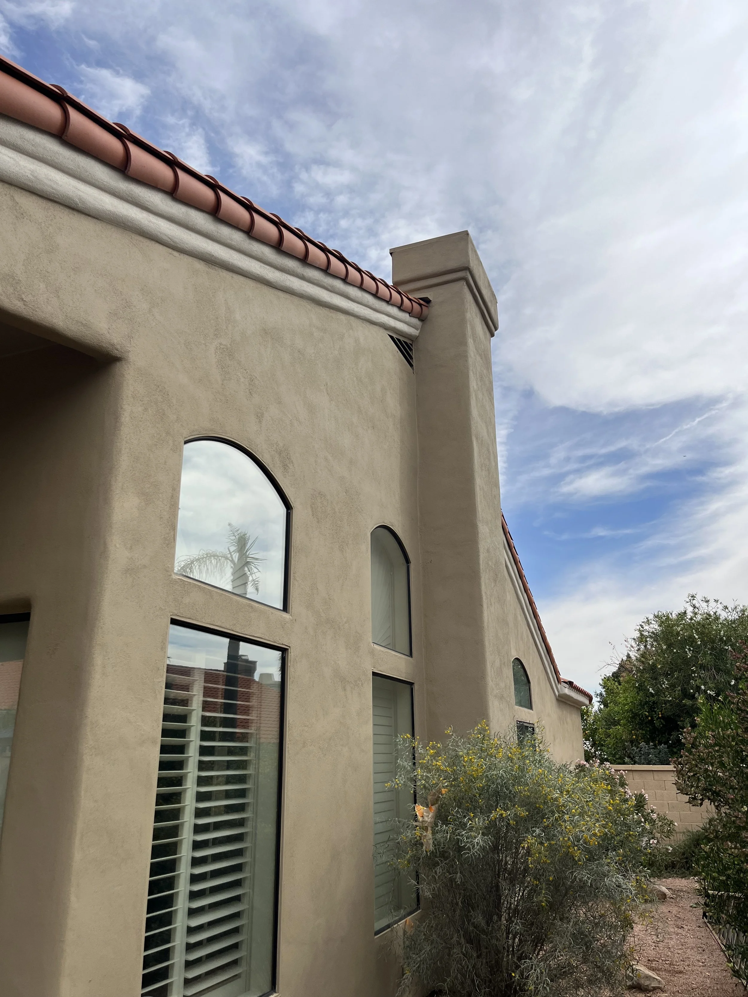 Side view of a beige stucco house with arched windows, red clay tile roof, chimney, and desert plants in the yard under a partly cloudy sky.
