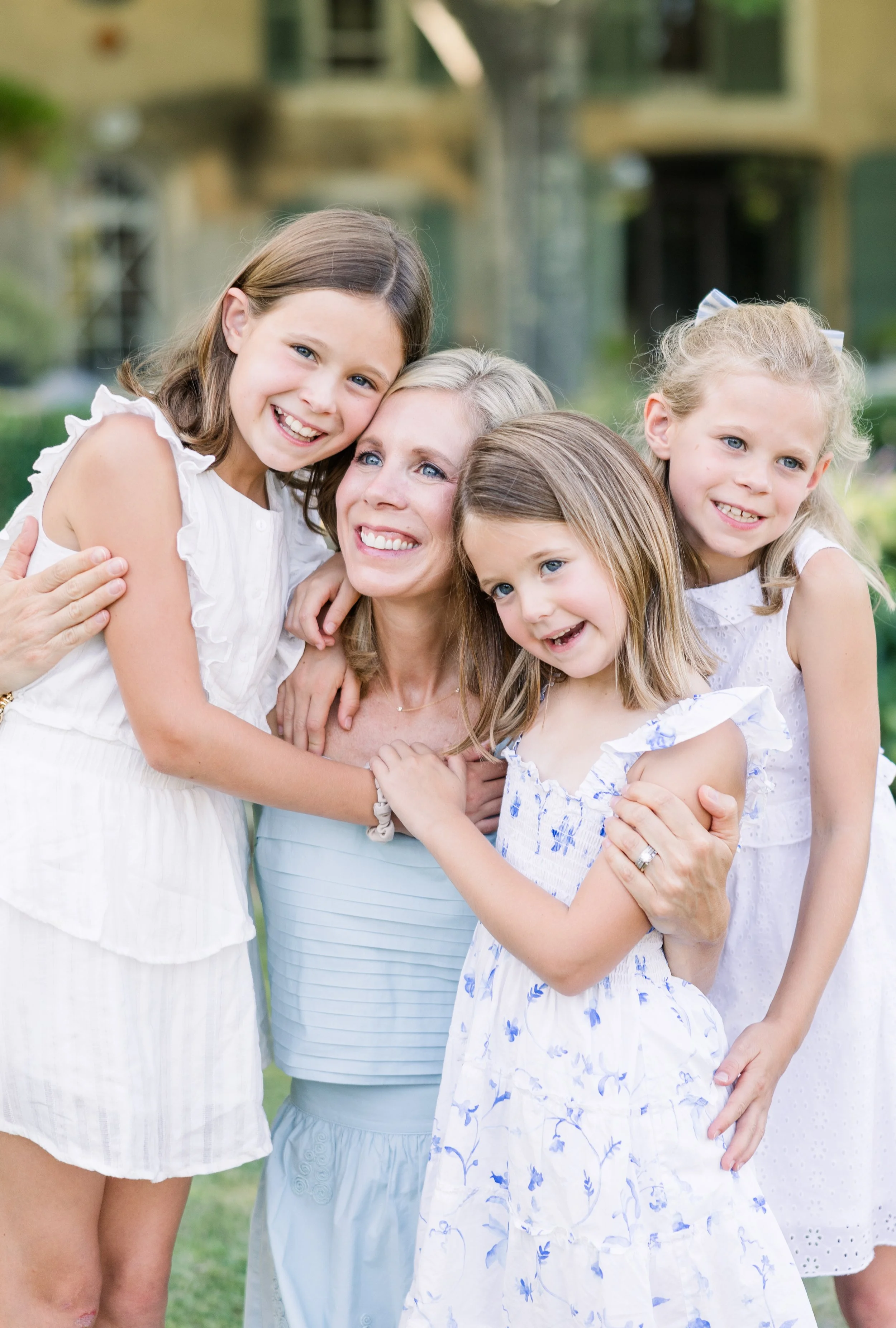 Mom hugging three smiling daughters in casual family photo with Provence villa green garden background