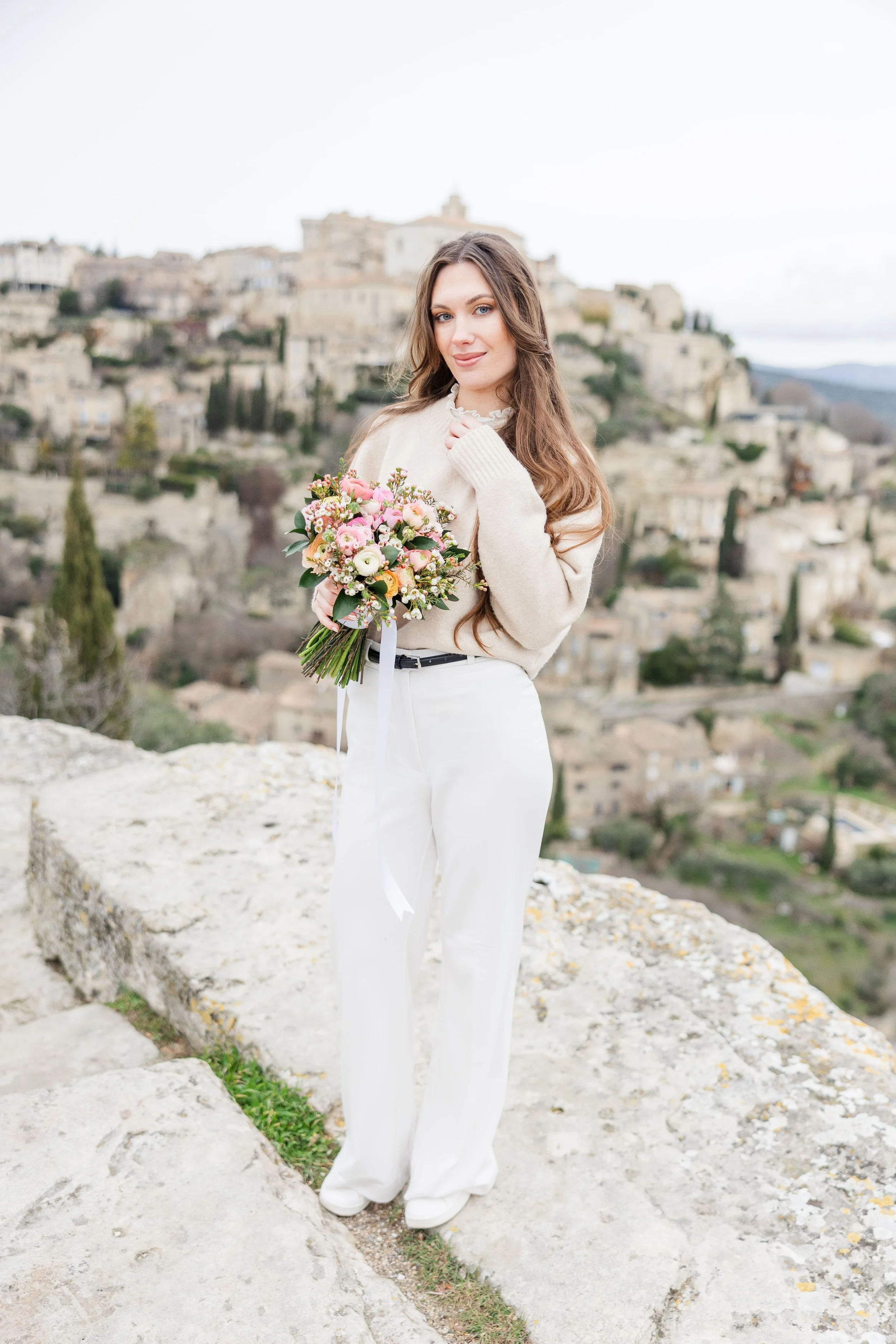 Woman holding a bouquet in front of the village of Gordes, Provence, captured in soft natural light during a romantic portrait session.