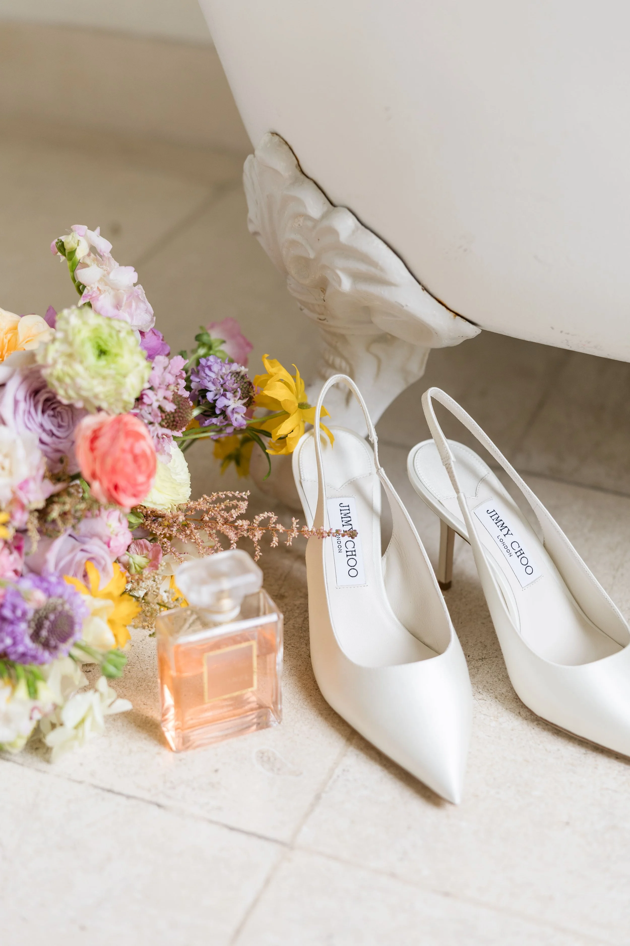 Wedding flat lay featuring Jimmy Choo bridal shoes, perfume, and pastel floral arrangements in soft blush and romantic tones.