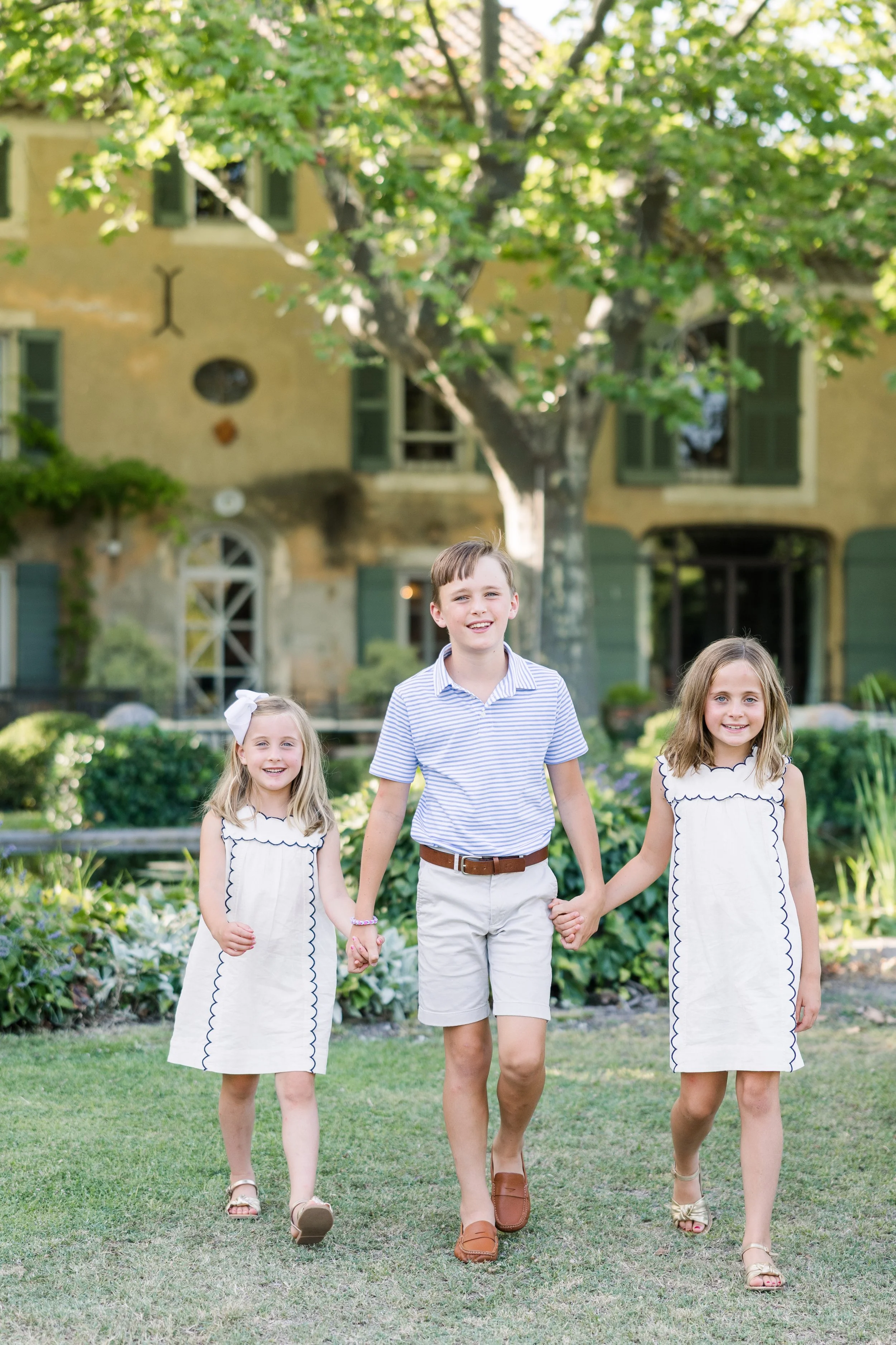 Three kids posing together outside a yellow Provence villa with green shutters and manicured garden