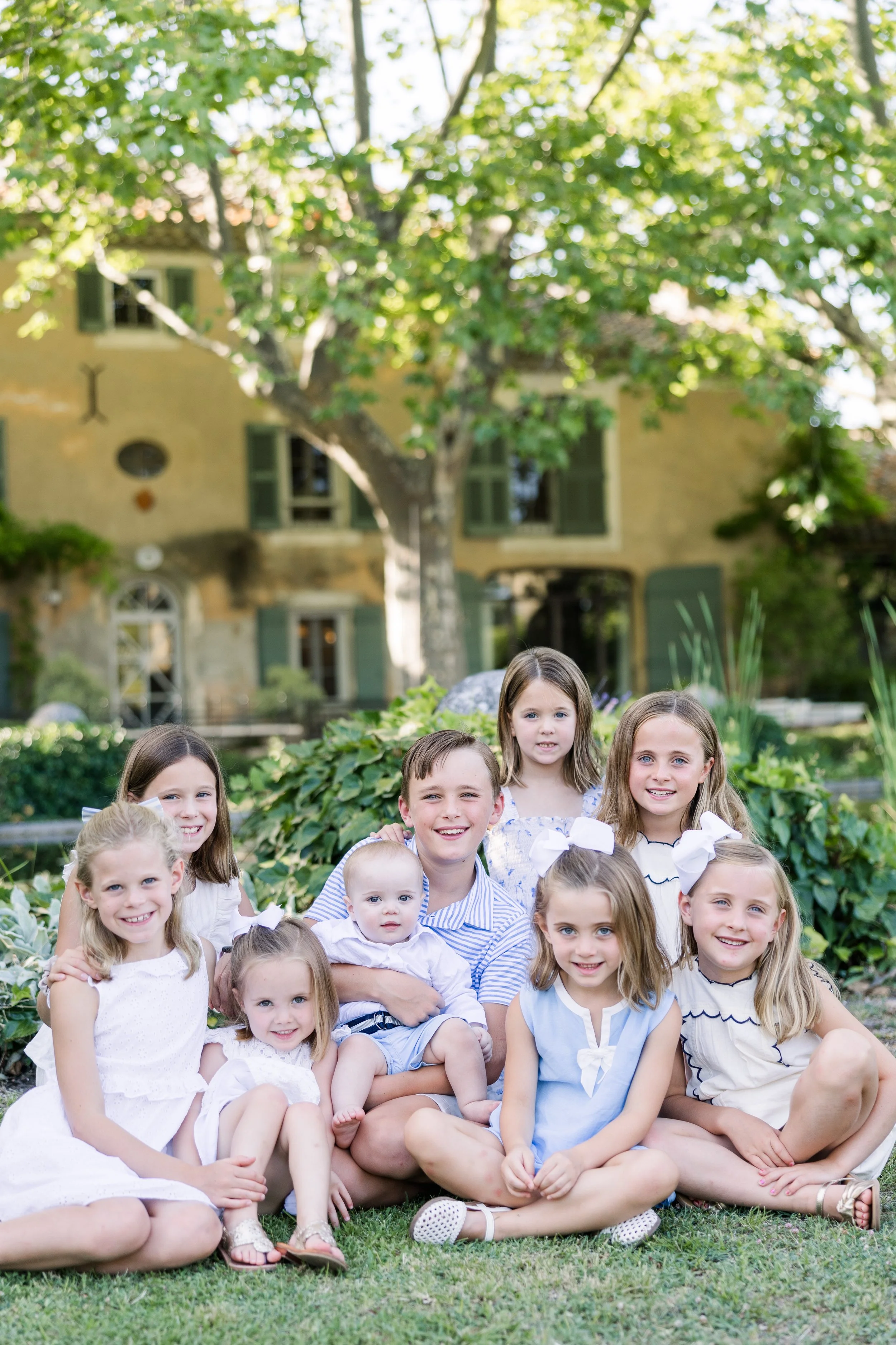 Family of night kids wearing neutral beige and white clothing standing together in front of tall trees at a Provence villa