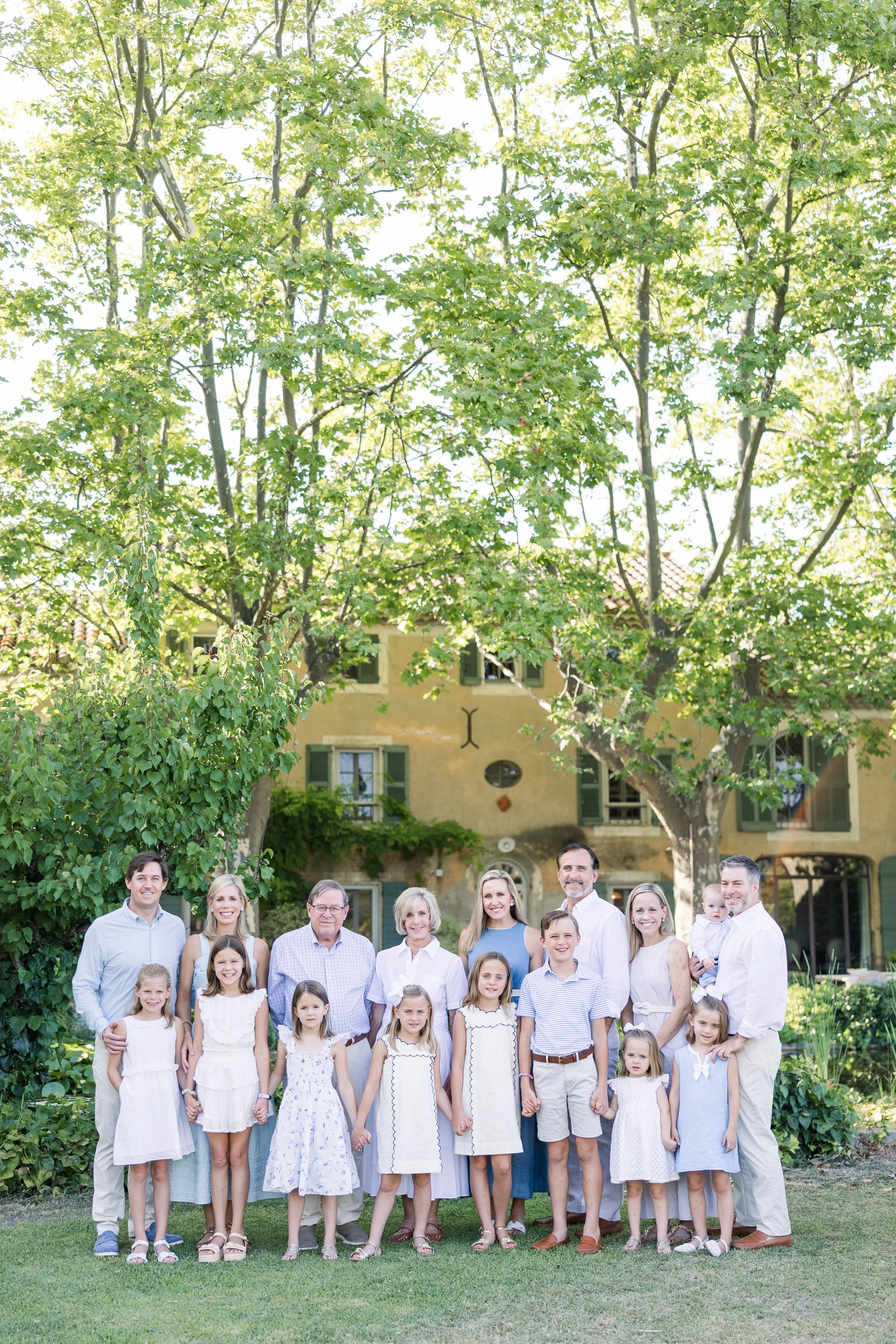 Large family group of ten people in light colored clothing outdoors in a Provence villa garden with mature trees