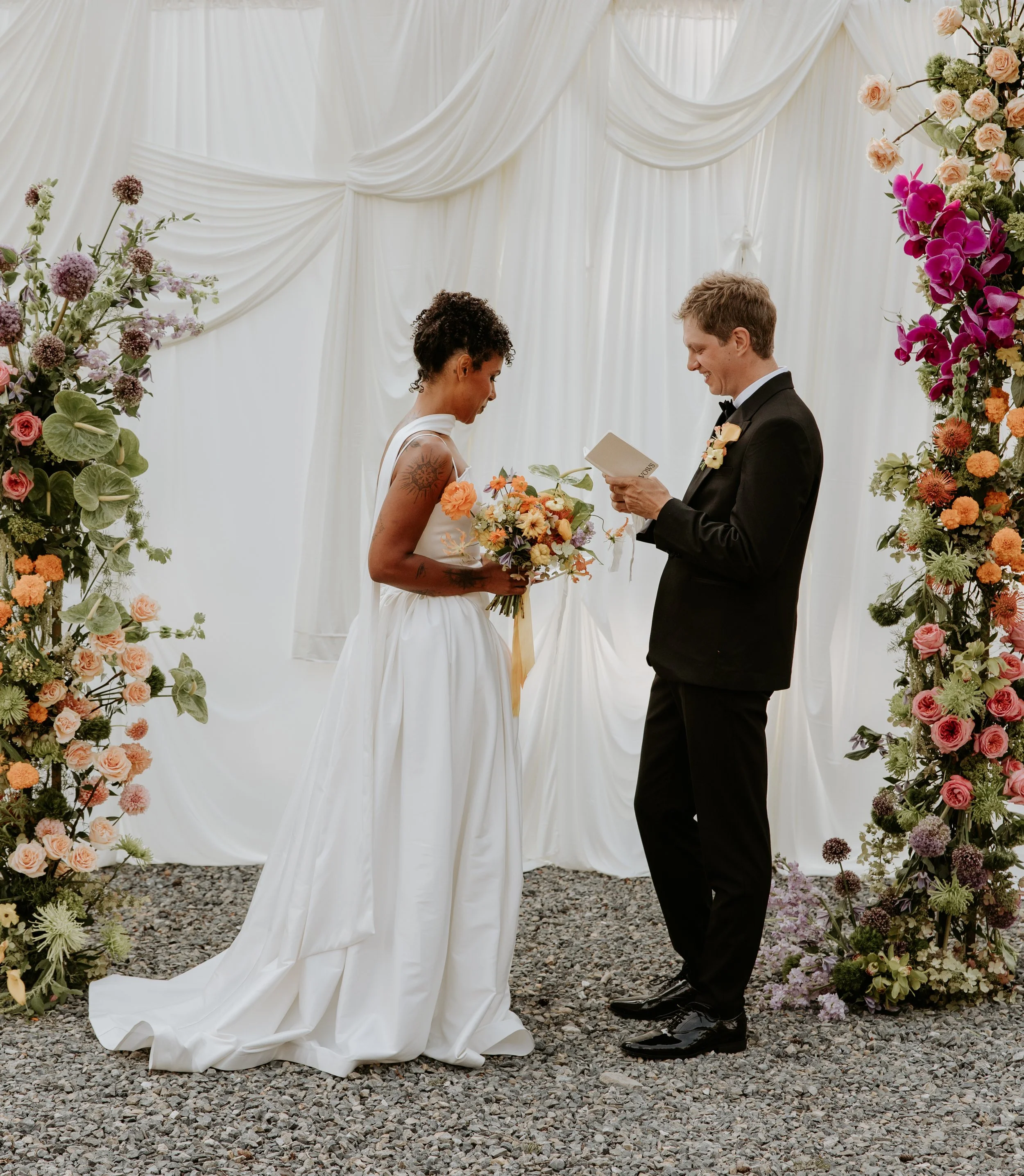 A couple at the altar reading vows at a Montana micro wedding. Woman is wearing a ballgown white dress, man in a black tux, floral arrangements on both sides with a white draped backdrop