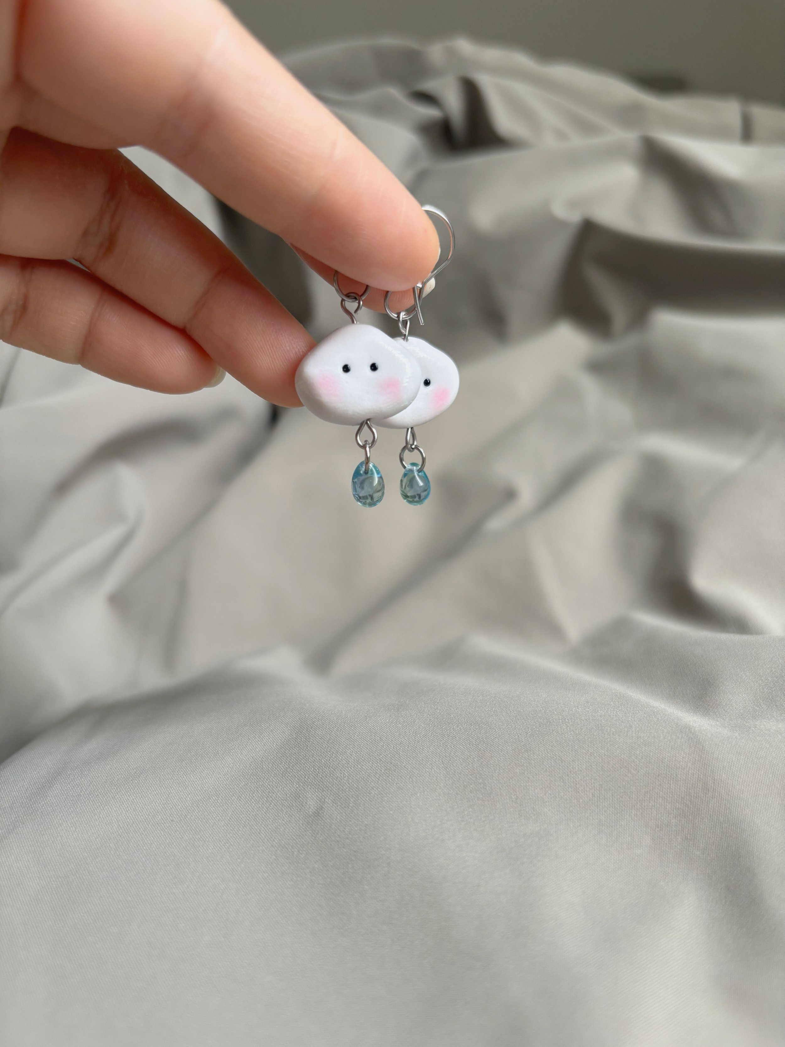 Close-up of a hand holding a pair of earrings with white cloud-shaped charms, pink blush on cheeks, black eyes, and small blue teardrop-shaped glass beads hanging below.