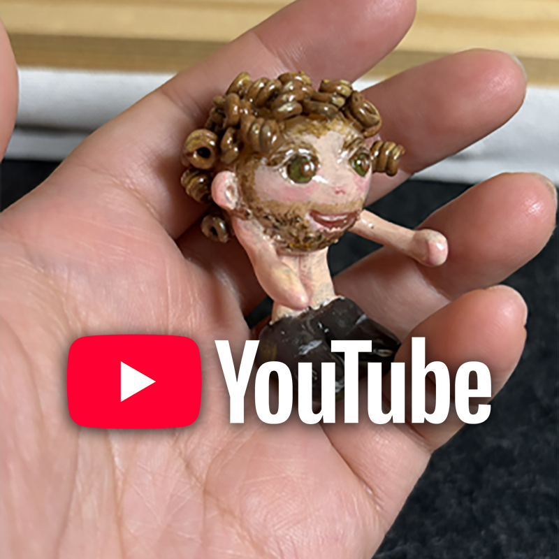 A hand holding a small painted figurine of a person with curly brown hair, a beard, and a smiling expression, with the YouTube logo overlaid. The image is linked to the CradlingCreative YouTube channel.