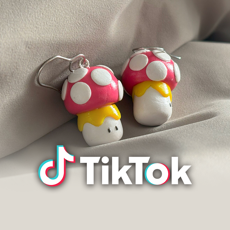 Pair of mushroom-shaped earrings with pink caps and white spots, on a soft fabric surface, with TikTok logo overlay. The image is linked to the CradlingCreative TikTok channel.