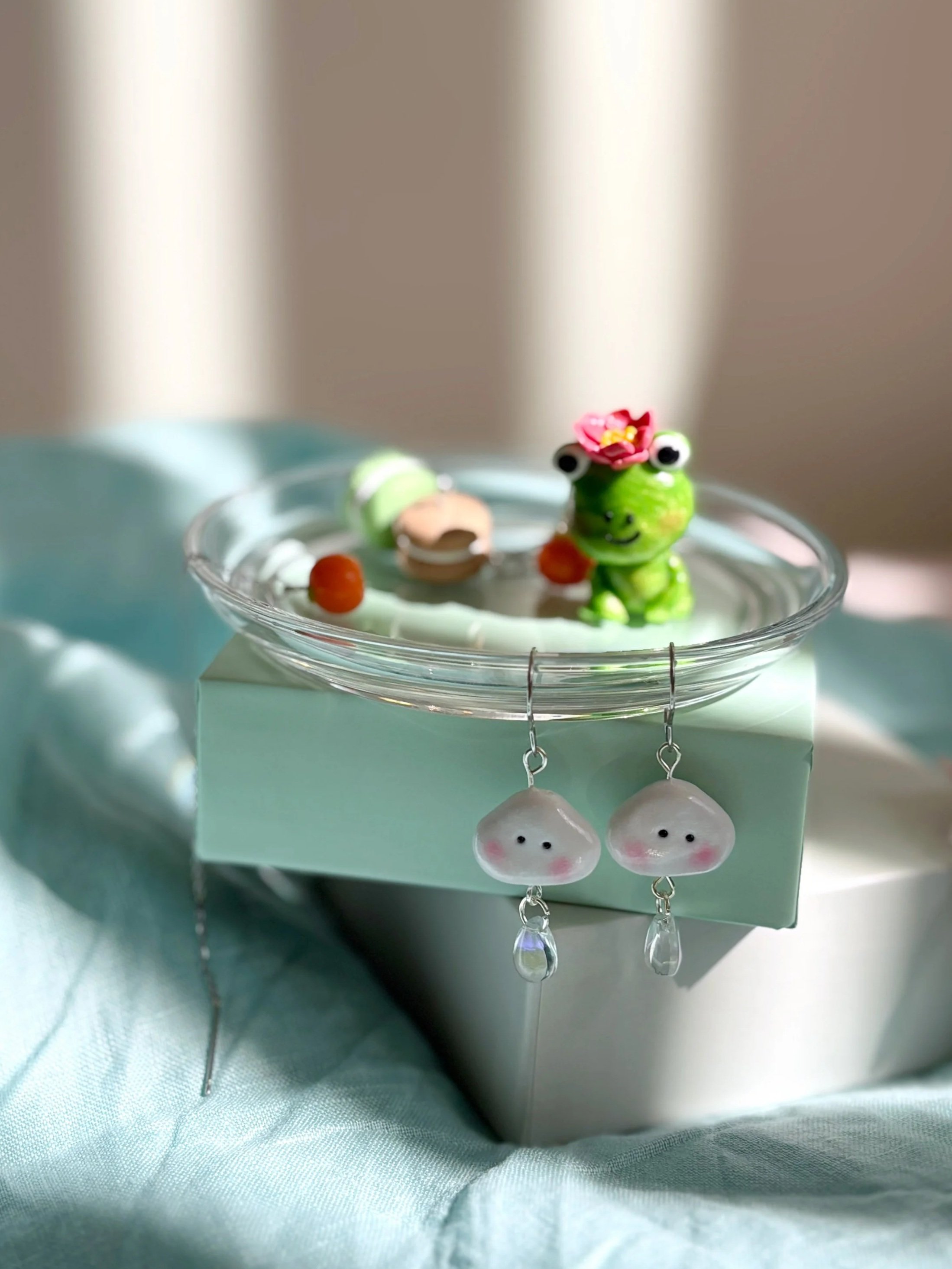 Decorative jewellery with a green frog figure wearing a pink flower on its head, placed in a glass dish with some earrings hanging on it, with background and fabric in shades of light blue and beige.