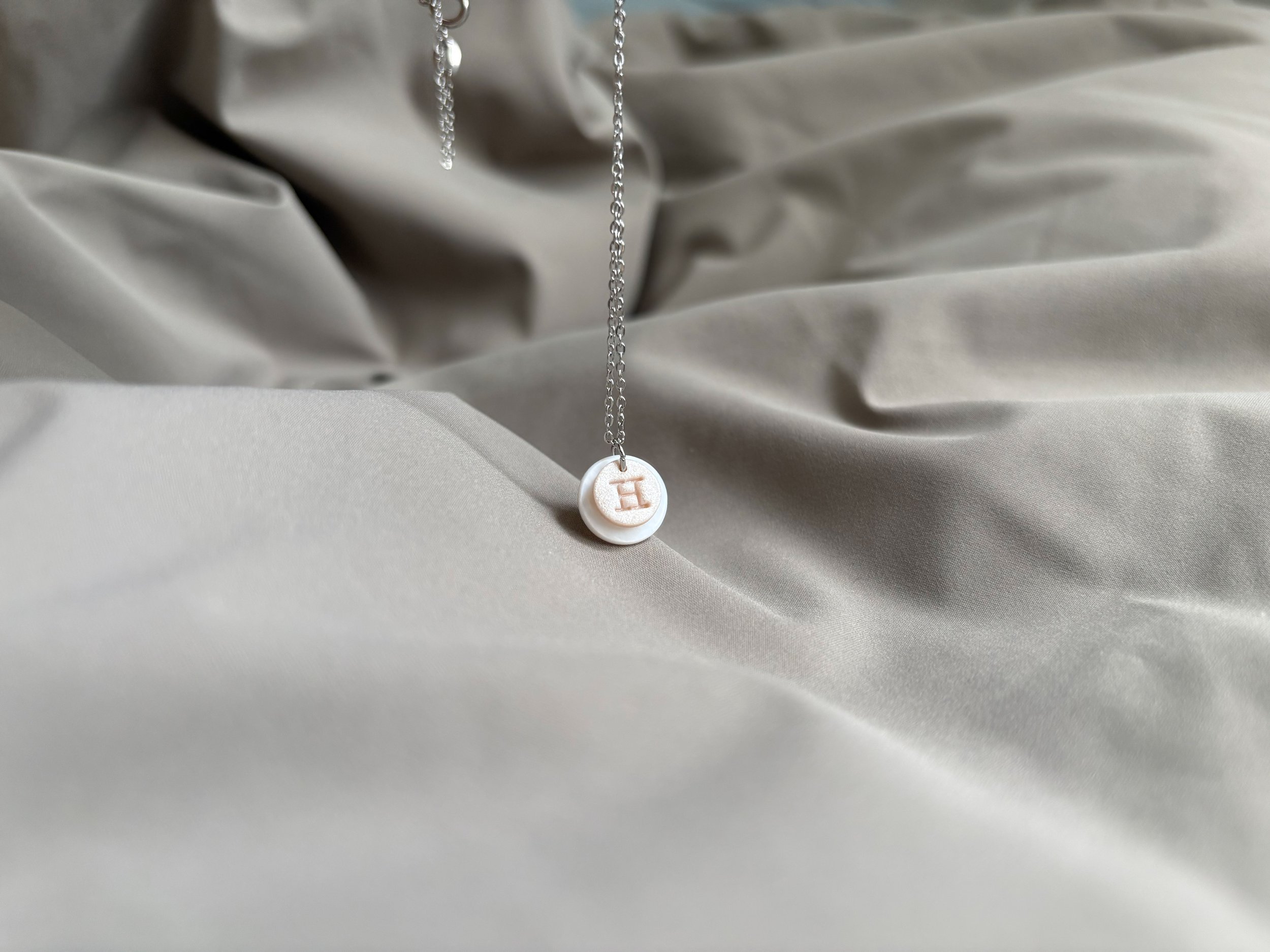 Silver necklace with a pendant featuring an initial letter 'H', hanging against a crinkled white satin background.