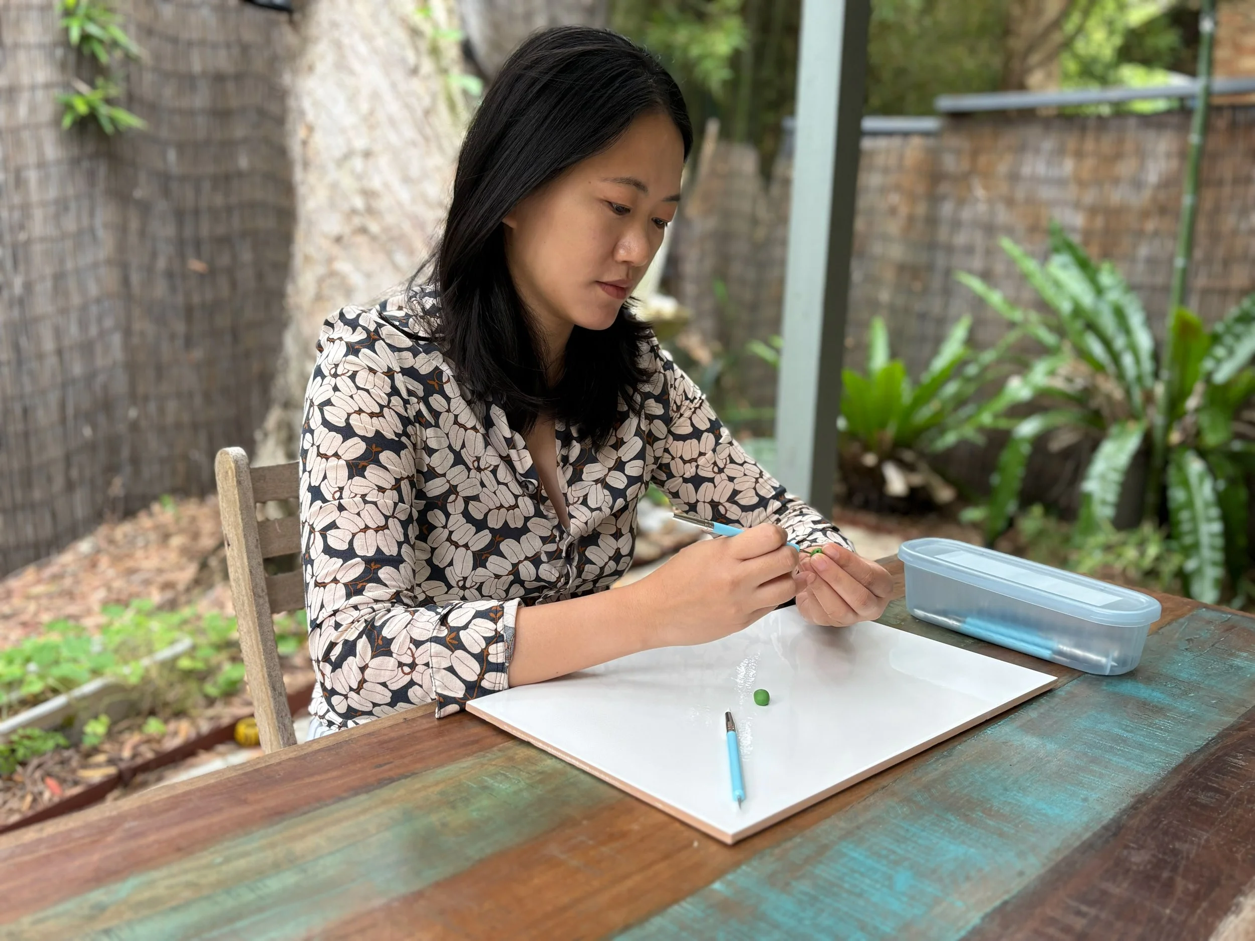 A young woman(founder of CradlingCreative) with black hair, wearing a patterned shirt, sitting at a wooden table outdoors, sculpting using polymer clay with various tools on the table.