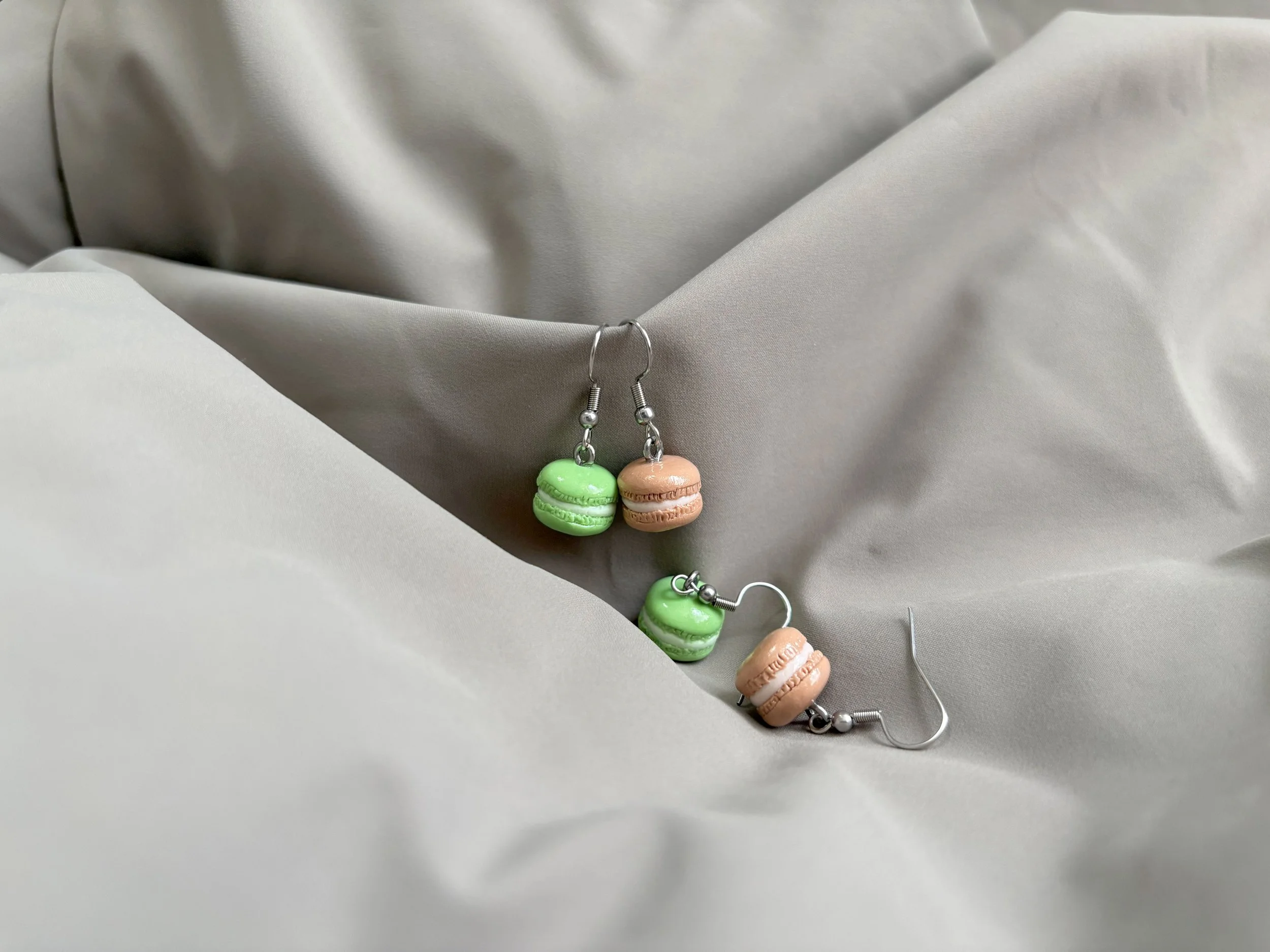 Two pairs of earrings featuring macarons, in both green and beige, with silver hoop earring hooks against a light grey, slightly wrinkled fabric background.