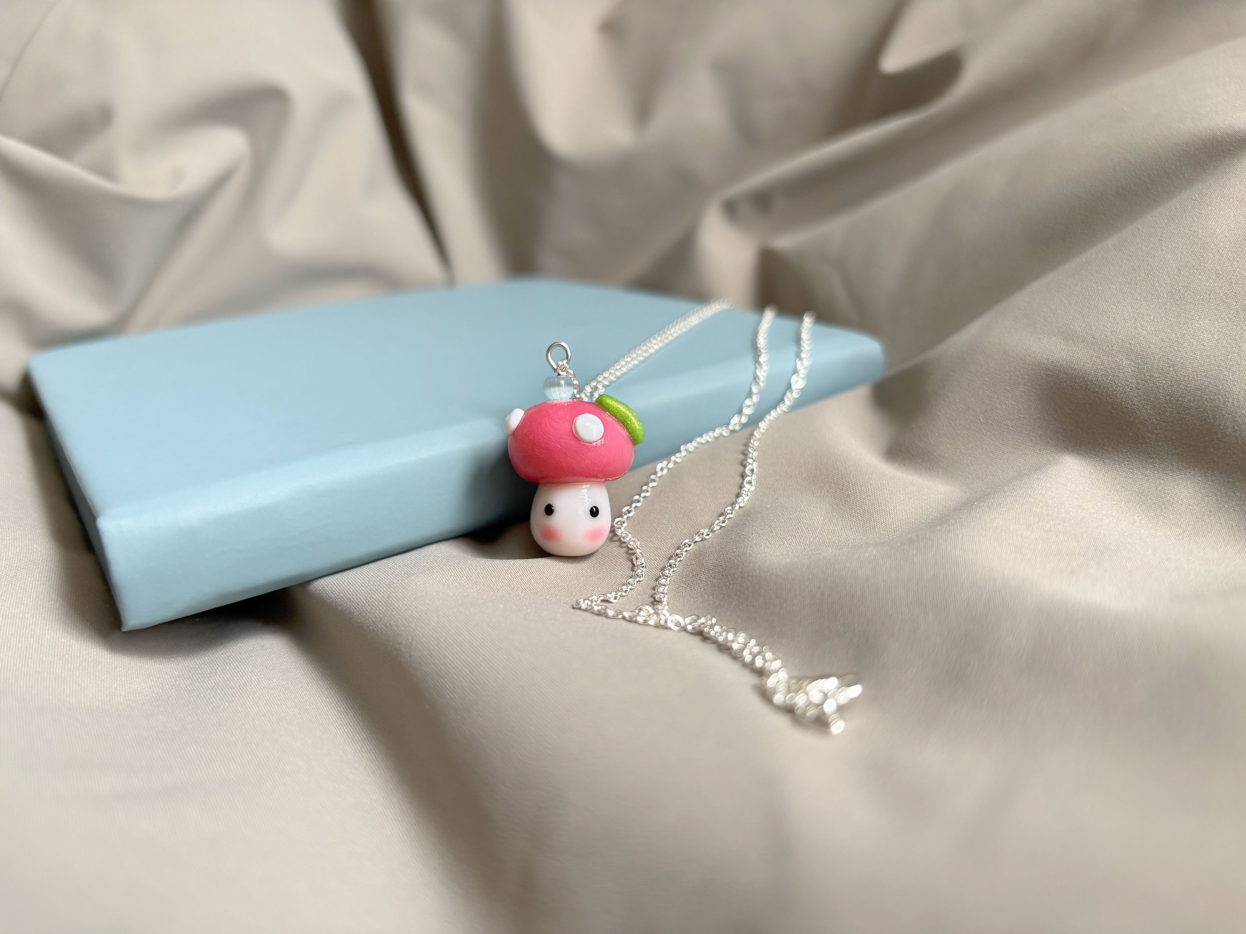 A necklace with a pendant resembling a small pink mushroom with white dots, hanging on a silver chain, was displayed on a light blue notebook with a satin fabric background.