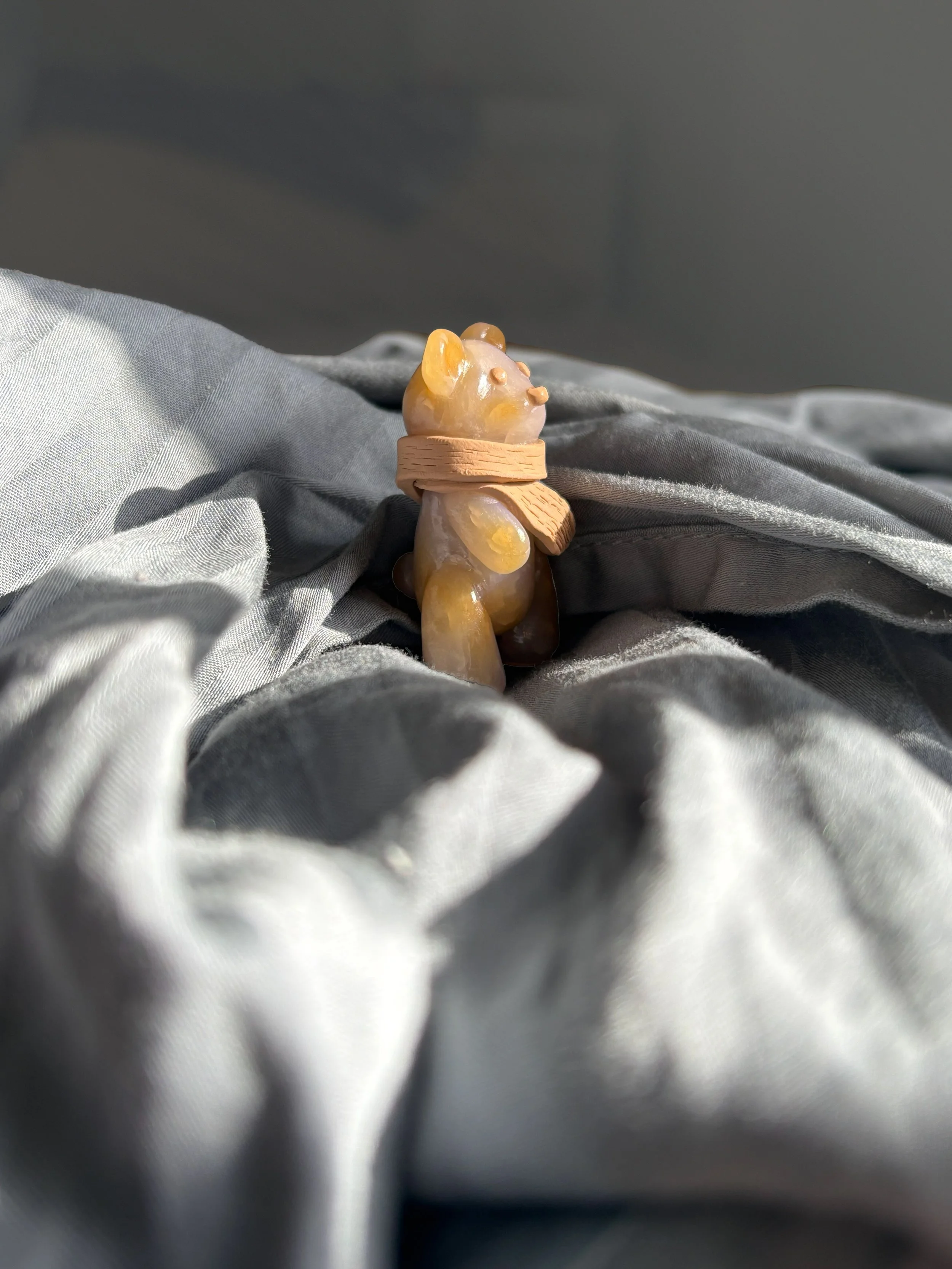 A small, bear-shaped figurine with yellow ears and paws, wrapped in a light-colored band or ribbon, sitting on a crumpled light gray fabric background.
