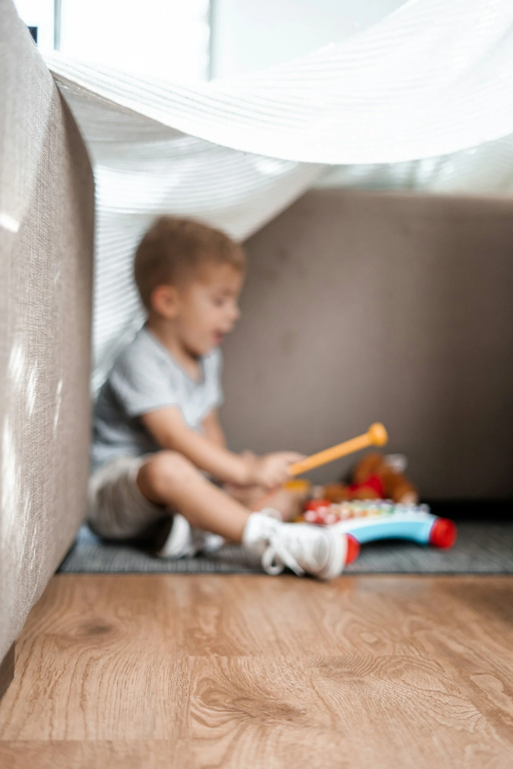 A young boy sitting on the floor under a bed, playing with toys. The photo is out of focus.
