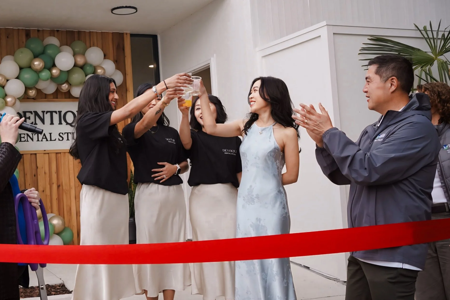 A group of people celebrating a special event, standing behind a red ribbon. Two women in black shirts and beige skirts are toasting with drinks, while a woman in a white dress and a man in a gray jacket are smiling and clapping. There is a balloon decoration and a sign that says 'Dentqi' in the background.