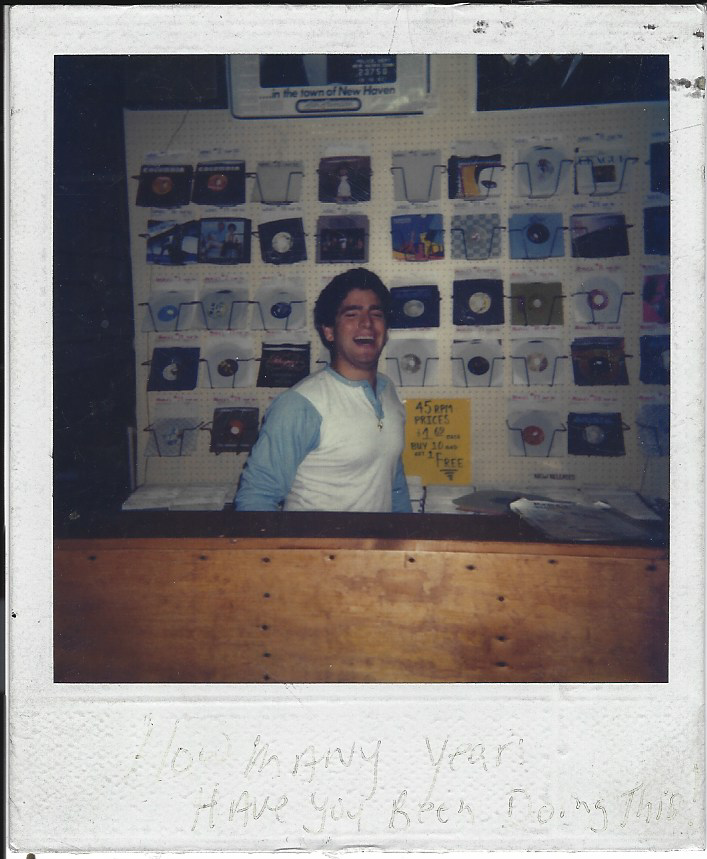 A young man selling records at a booth, smiling and wearing a light blue and white raglan shirt, with a sign indicating discounted prices for 45 RPM records in the background.