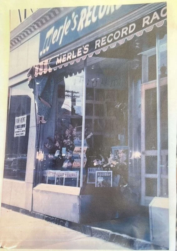 A vintage storefront with a sign reading 'Tops Records' and a awning that says 'Merle's Record Racket.' There is a window display with flowers and records, and a 'For Rent' sign on the door.