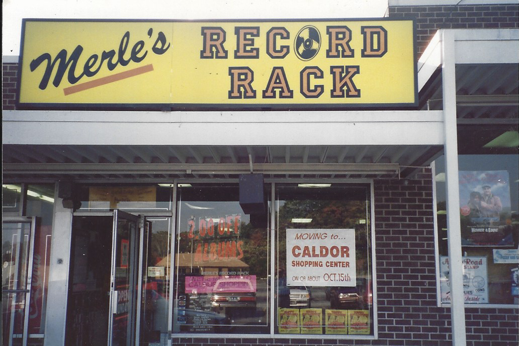 Storefront sign for Merle's Record Rack, with a notice about moving to Caldor Shopping Center on October 15th, and the store's glass door and window showing reflections and posters.