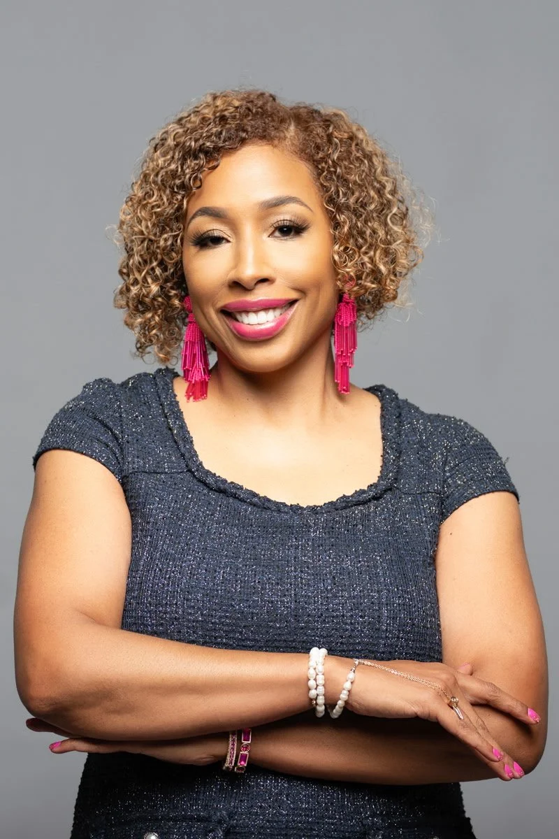 A woman with curly blonde hair, wearing pink earrings and a dark sparkly top, smiling with her arms crossed against a gray background.