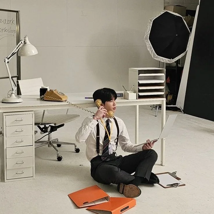 A young man in office attire sitting on the floor in a photography studio, holding a vintage rotary phone to his ear with one hand and a laptop in the other. The studio background includes a white desk, a white office chair, a large studio umbrella,