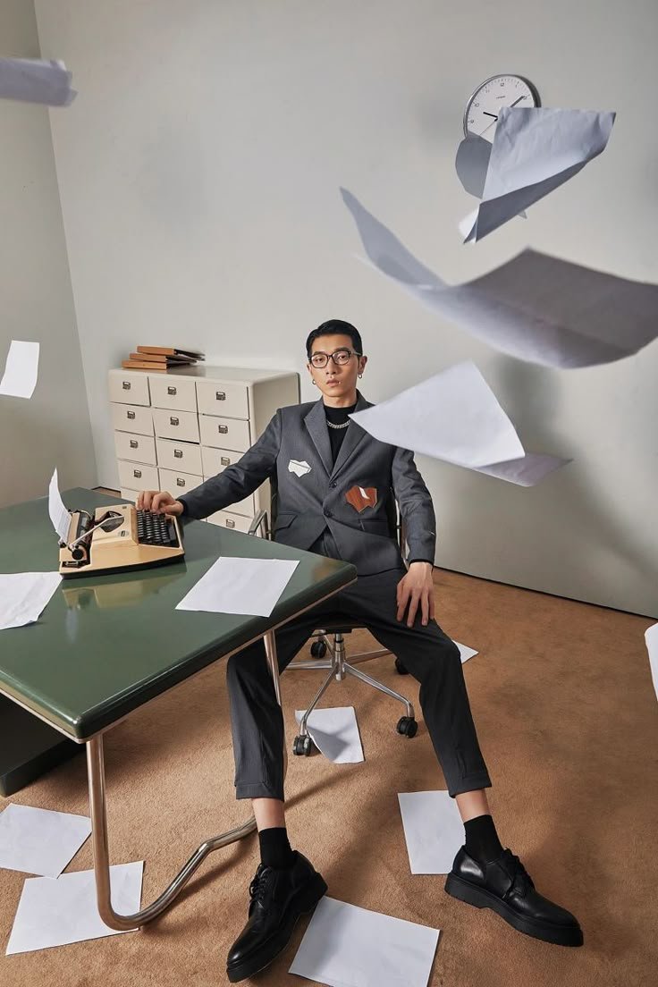 A person wearing glasses in a gray suit sits at a desk with scattered papers flying around the room, with a white wall and a clock in the background.
