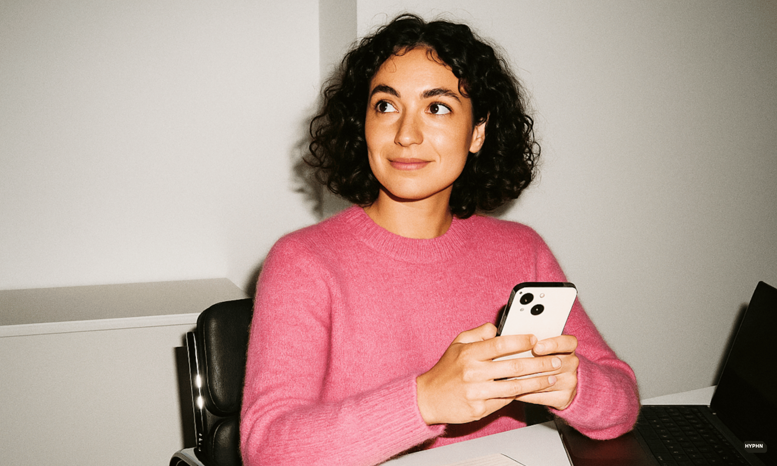 Woman wearing a bright pink jumper sits at a desk holding her mobile phone with a slight smile. 