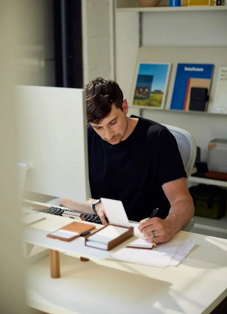 male employee in a black t-shirt sits at his desk in an office and writes notes in front of his computer.