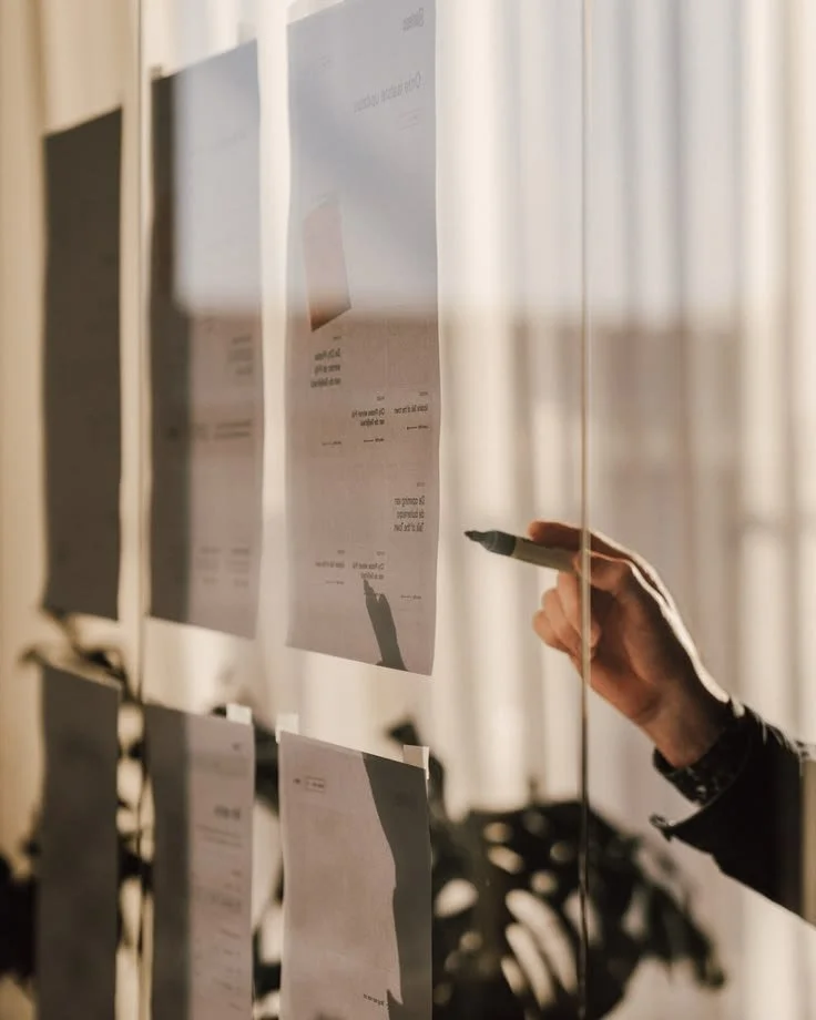 A person's hand holding a marker, pointing at a paper attached to a vertical glass board with other papers, in a well-lit room with curtains in the background.