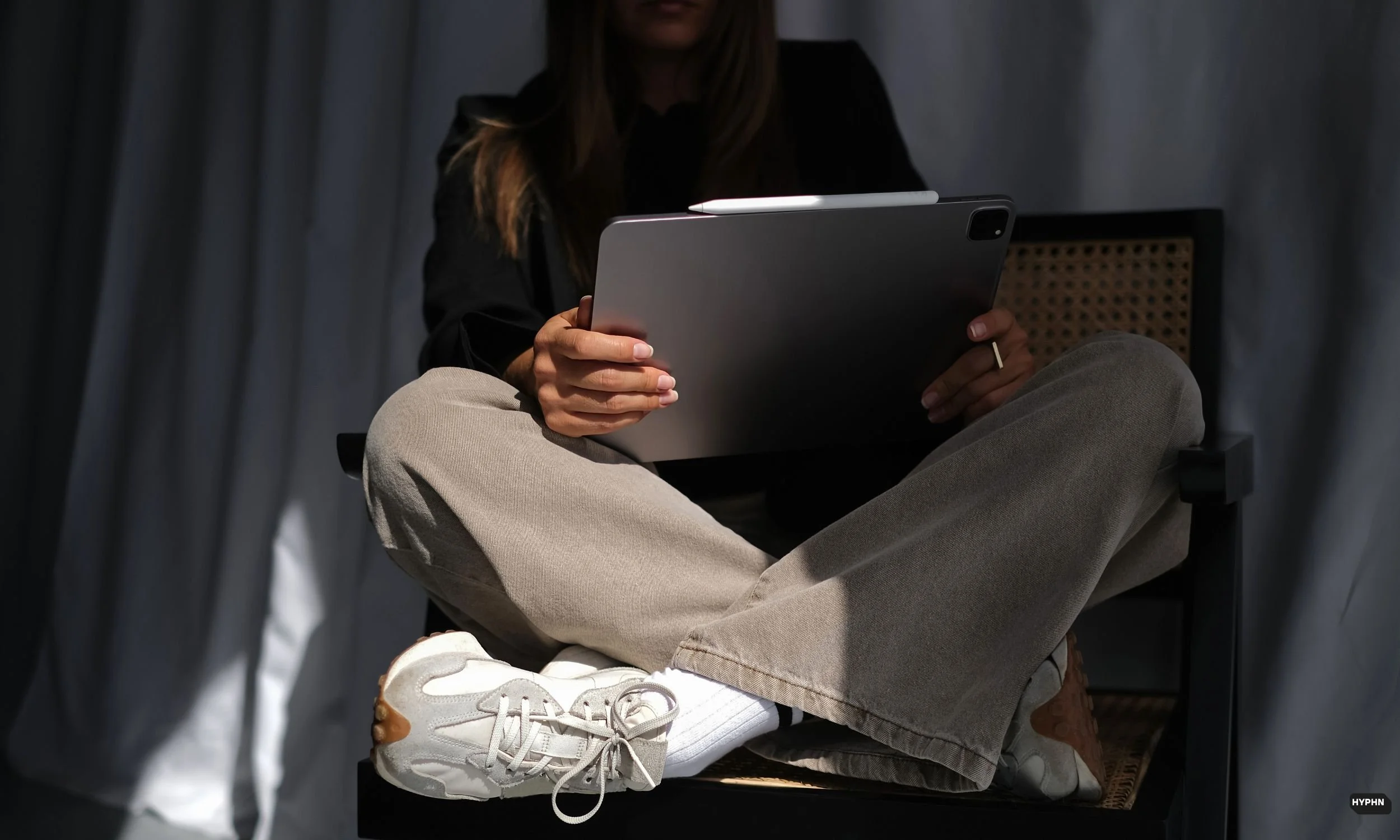 A person sitting cross-legged on a chair, holding a tablet device, with gray pants, white sneakers, and a black top, in front of gray curtains.